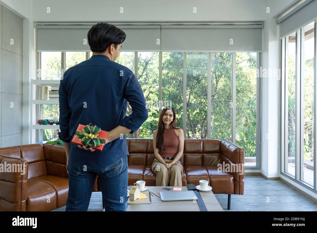 Les jeunes couples passent des vacances dans le salon. Le jeune homme porte des vêtements confortables, cache une boîte cadeau derrière, se préparant à surprendre la petite amie Banque D'Images