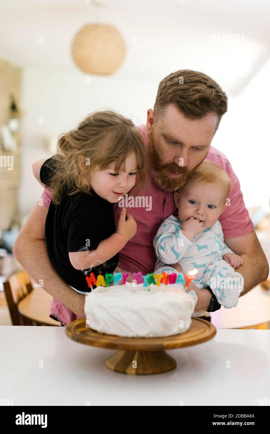 Père transportant des enfants (2-3, 6-11 mois) et des bougies d'anniversaire soufflantes sur le gâteau Banque D'Images