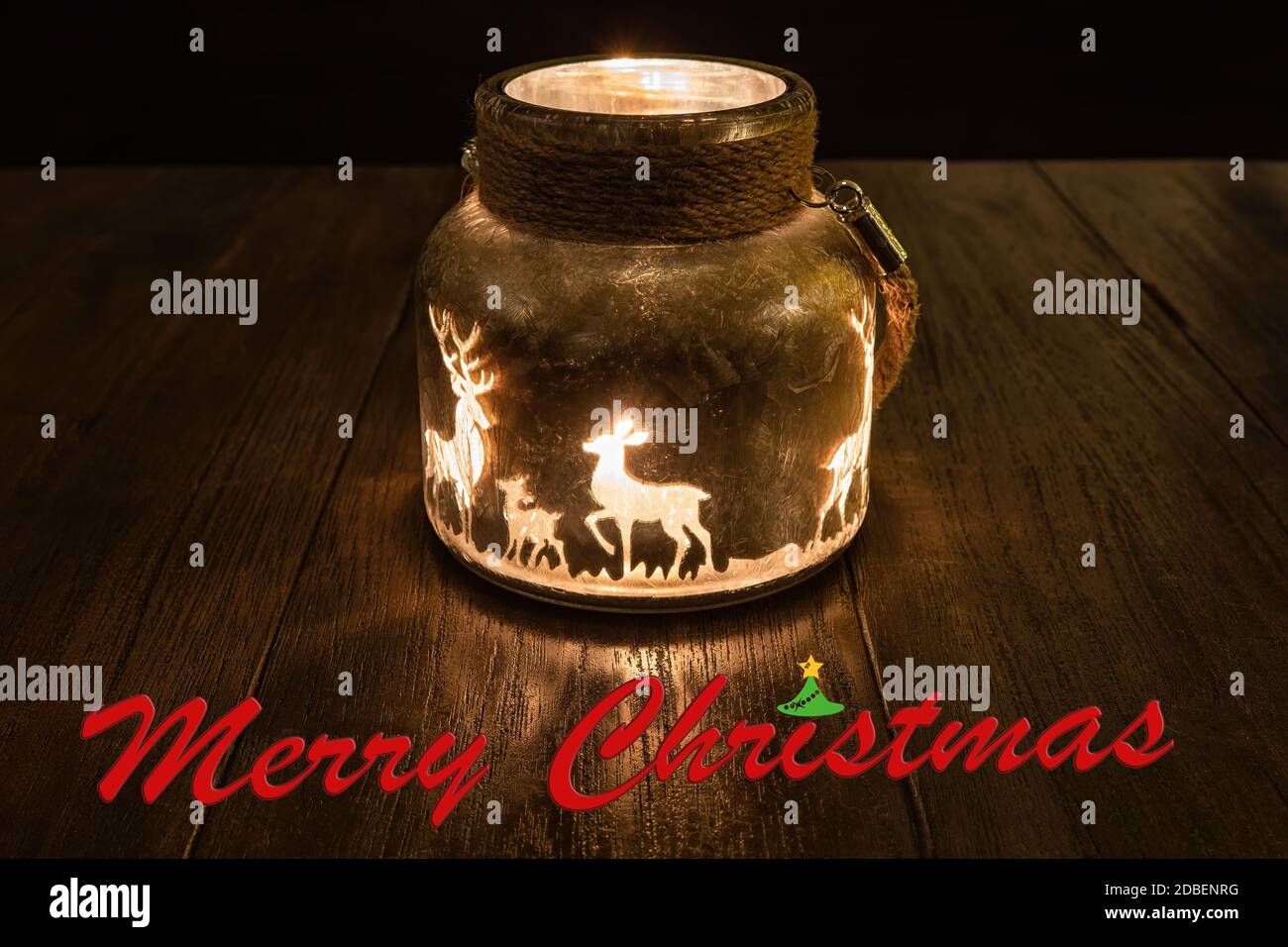Un verre de bougie brillant dérivé dans l'obscurité avec renne dessus et le lettrage Joyeux Noël au premier plan Banque D'Images