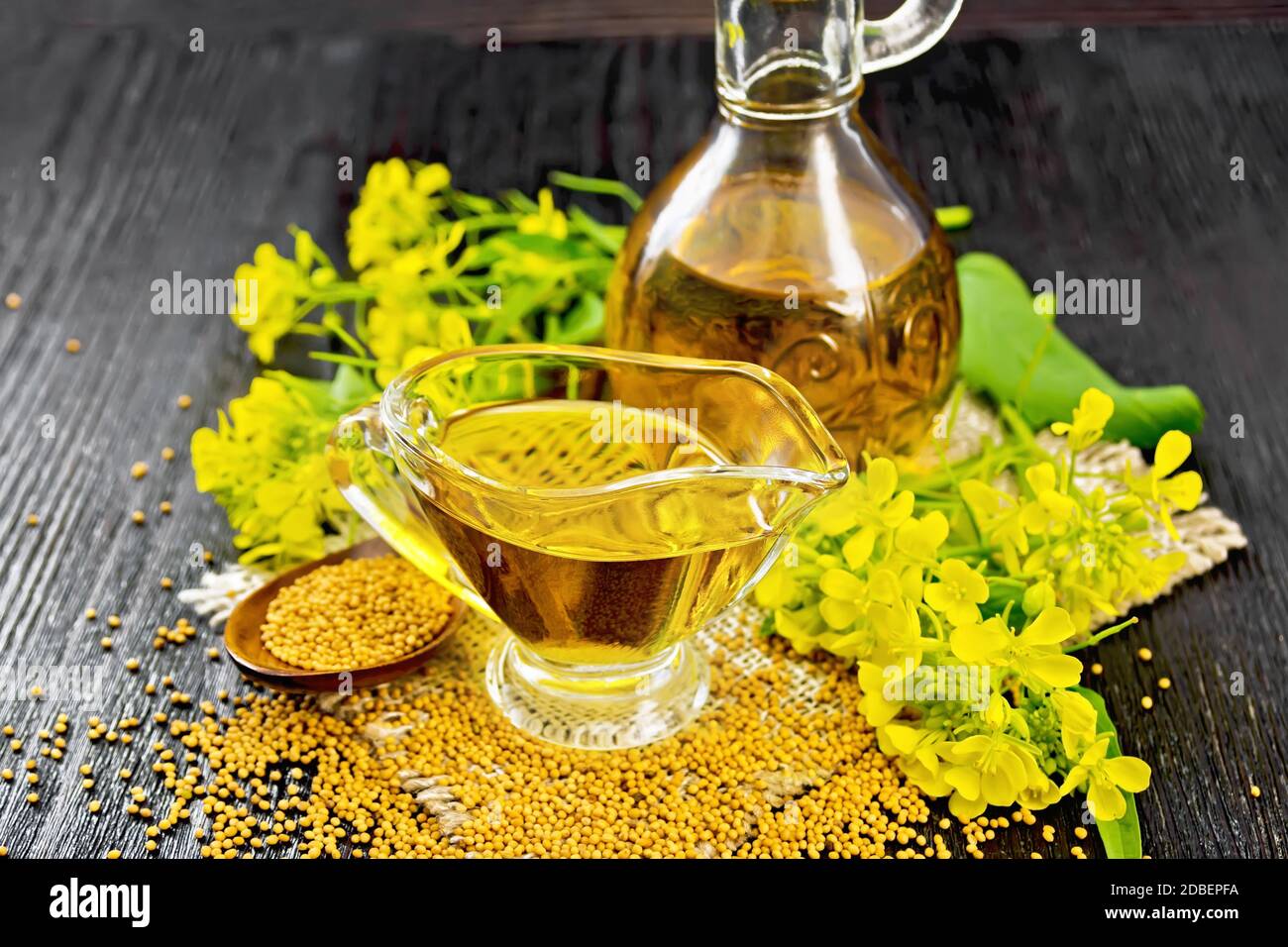 Huile de moutarde dans un bateau à sauce en verre et un décanteur, grains dans une cuillère, feuilles et fleurs de moutarde jaune sur le burap contre le panneau en bois sombre Banque D'Images