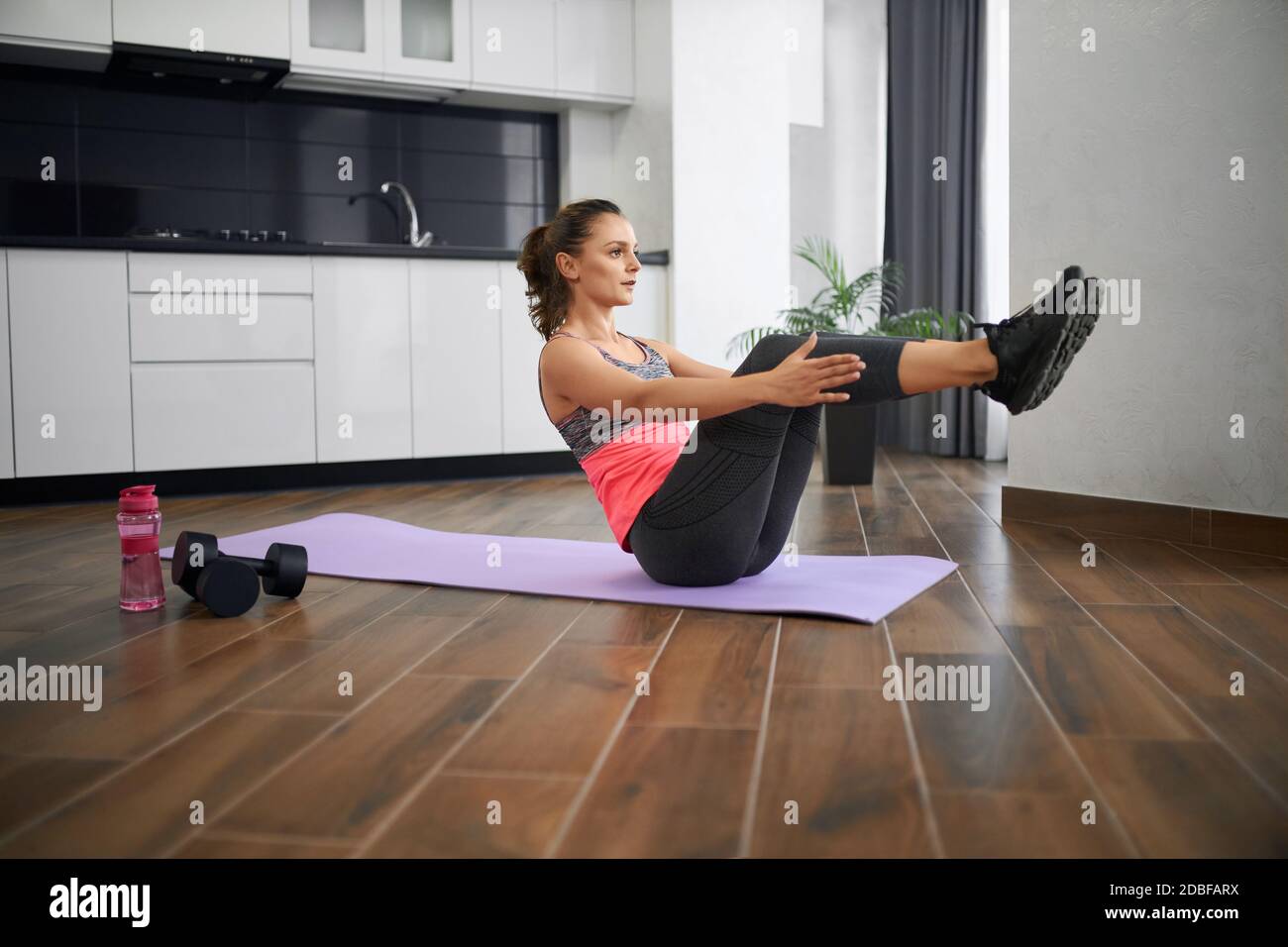 Vue latérale d'une jeune femme forte pratiquant la pose statique dans la cuisine. FIT fille portant des vêtements de sport entraînement abs, assis sur le tapis à la maison le matin. Concept d'entraînement à domicile, sport. Banque D'Images