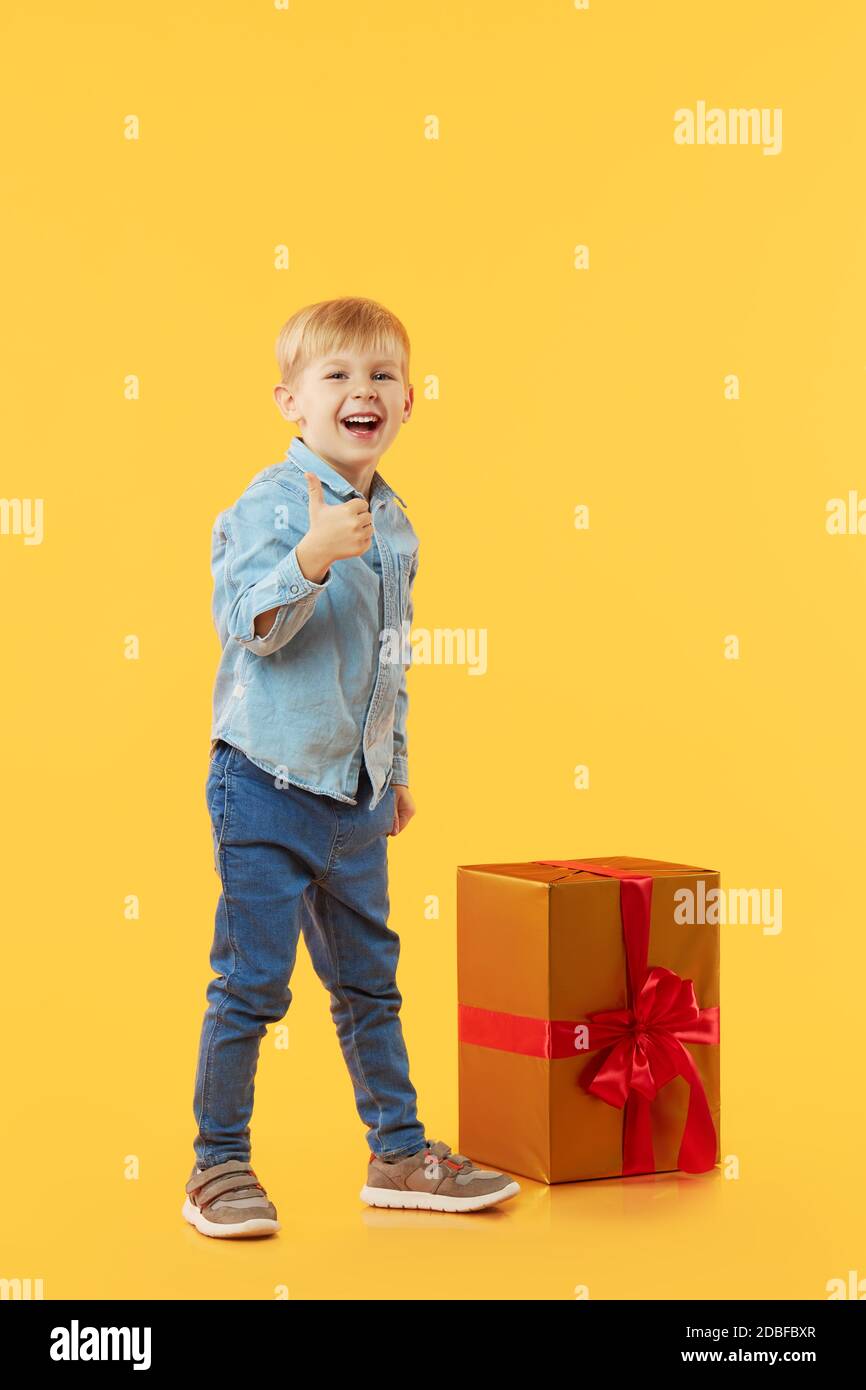 Portrait de l'enfant de smiling satisfait garçon avec grande boîte cadeau dorée montre pouce vers le haut, ok signe et regardant l'appareil photo sur fond jaune. Visage amusant Banque D'Images