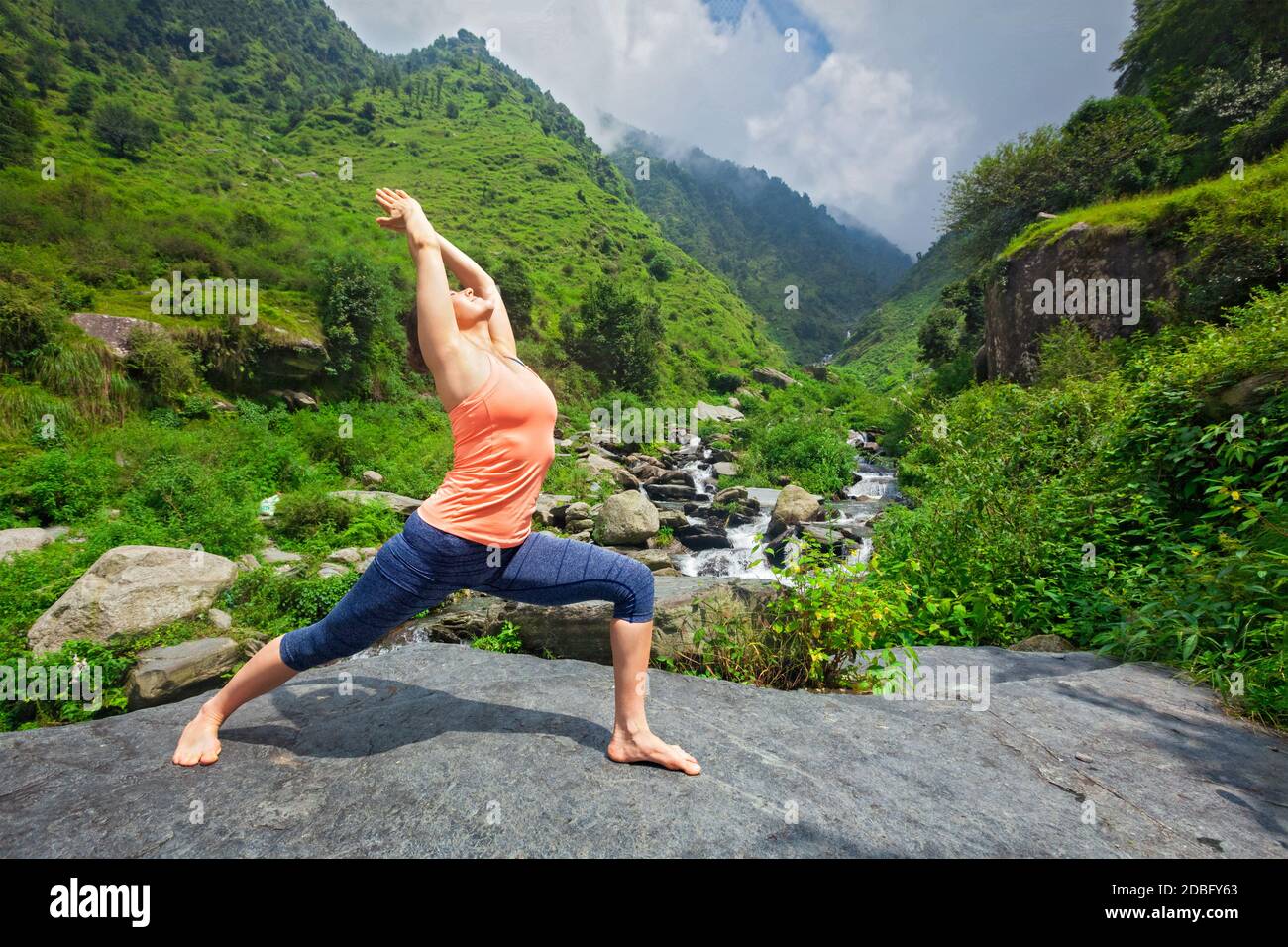 Yoga à l'extérieur - femme sportive faisant Ashtanga Vinyasa Yoga asana Virabhadrasana 1 Warrior pose la posture à la cascade dans l'Himalaya montagnes Banque D'Images