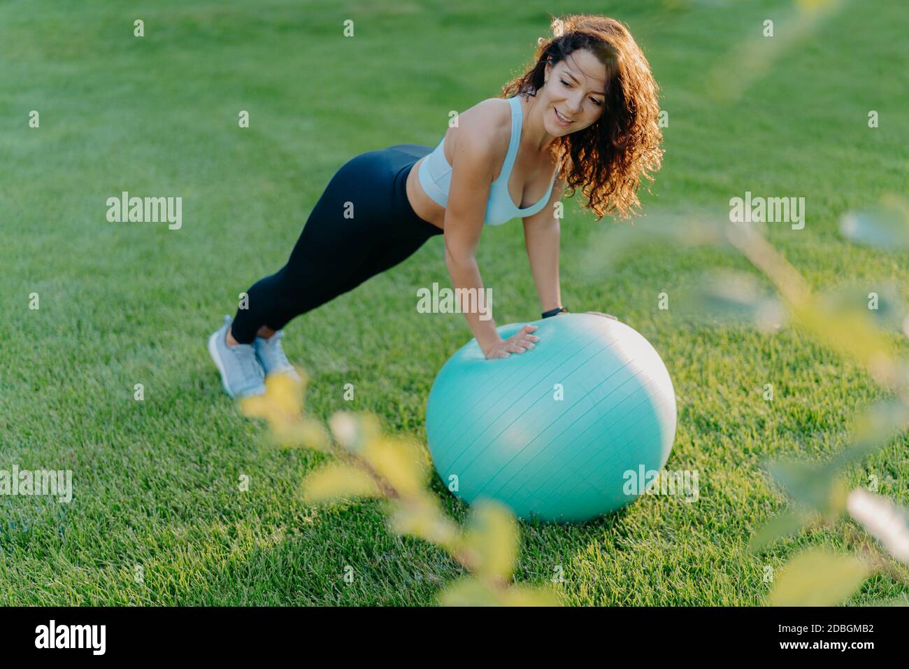 Femme ravie mince se penche au ballon d'exercice, vêtue de vêtements actifs, a des exercices de gymnastique sur la pelouse verte, aime le sport et l'activité préférée, mène en bonne santé Banque D'Images