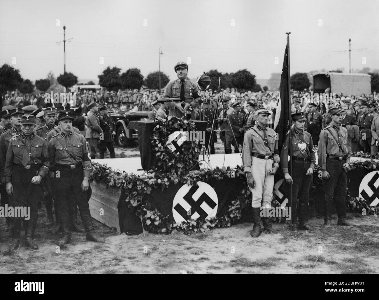 Au Congrès du Parti nazi de Nuremberg, un fonctionnaire du NSDAP prononce un discours sur un podium orné de drapeaux de la croix gammée. Derrière lui se cache une automobile. Banque D'Images