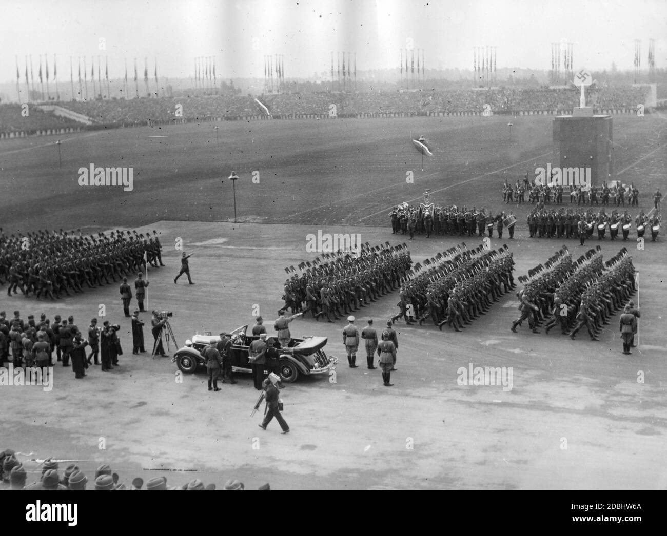 Adolf Hitler, dans sa Mercedes, prend le salut des formations du Service du travail de Reich sur le champ de Zeppelin. À sa gauche, un caméraman filme la scène. En arrière-plan, il y a le Mémorial du travail. Devant elle il y a un groupe de cuivres et un groupe de batteurs. Banque D'Images