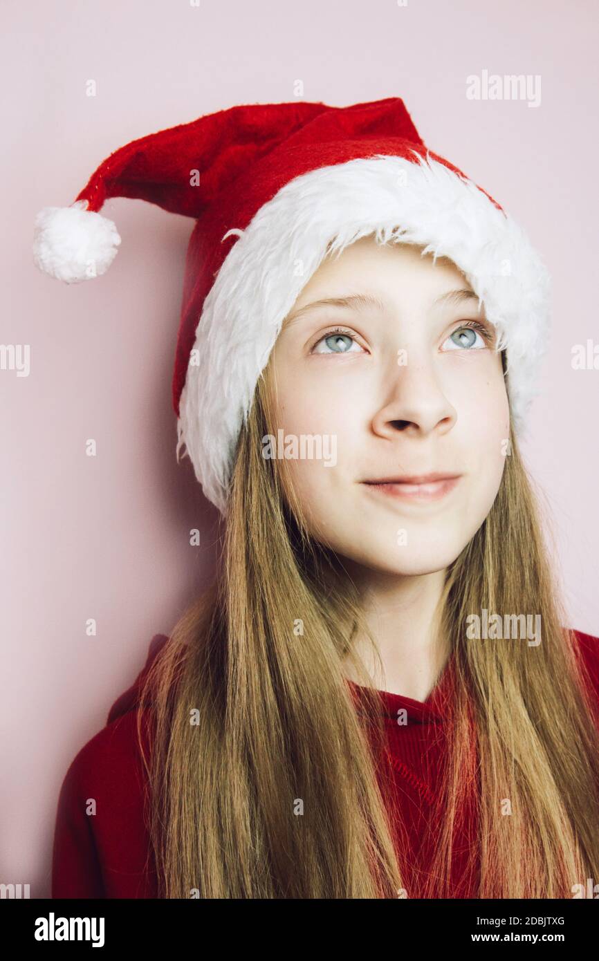 Belle fille souriante avec de longs cheveux blonds dans un chapeau de Père Noël moelleux sur fond rose. Adolescent aux émotions vives. Gros plan Banque D'Images