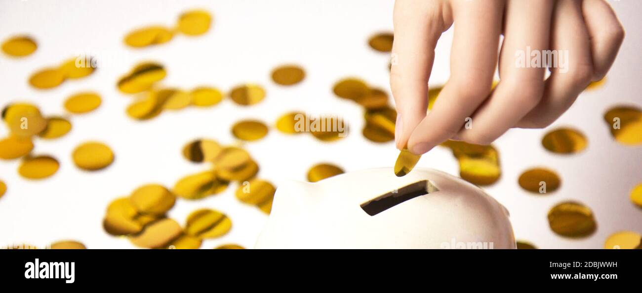 Mettez la pièce à la main à la tirelire. Banc de porc blanc sur fond de confetti de papier doré isolé sur fond blanc. Concept d'économie d'argent Banque D'Images