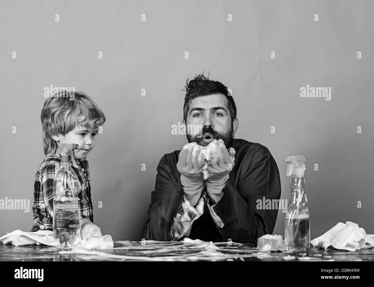 Homme avec la barbe et la moustache avec des gants en caoutchouc soufflant sur la mousse dans ses mains. Papa et son fils sur fond vert. Homme et enfant près de la table avec fournitures de nettoyage. Concept de temps familial. Banque D'Images