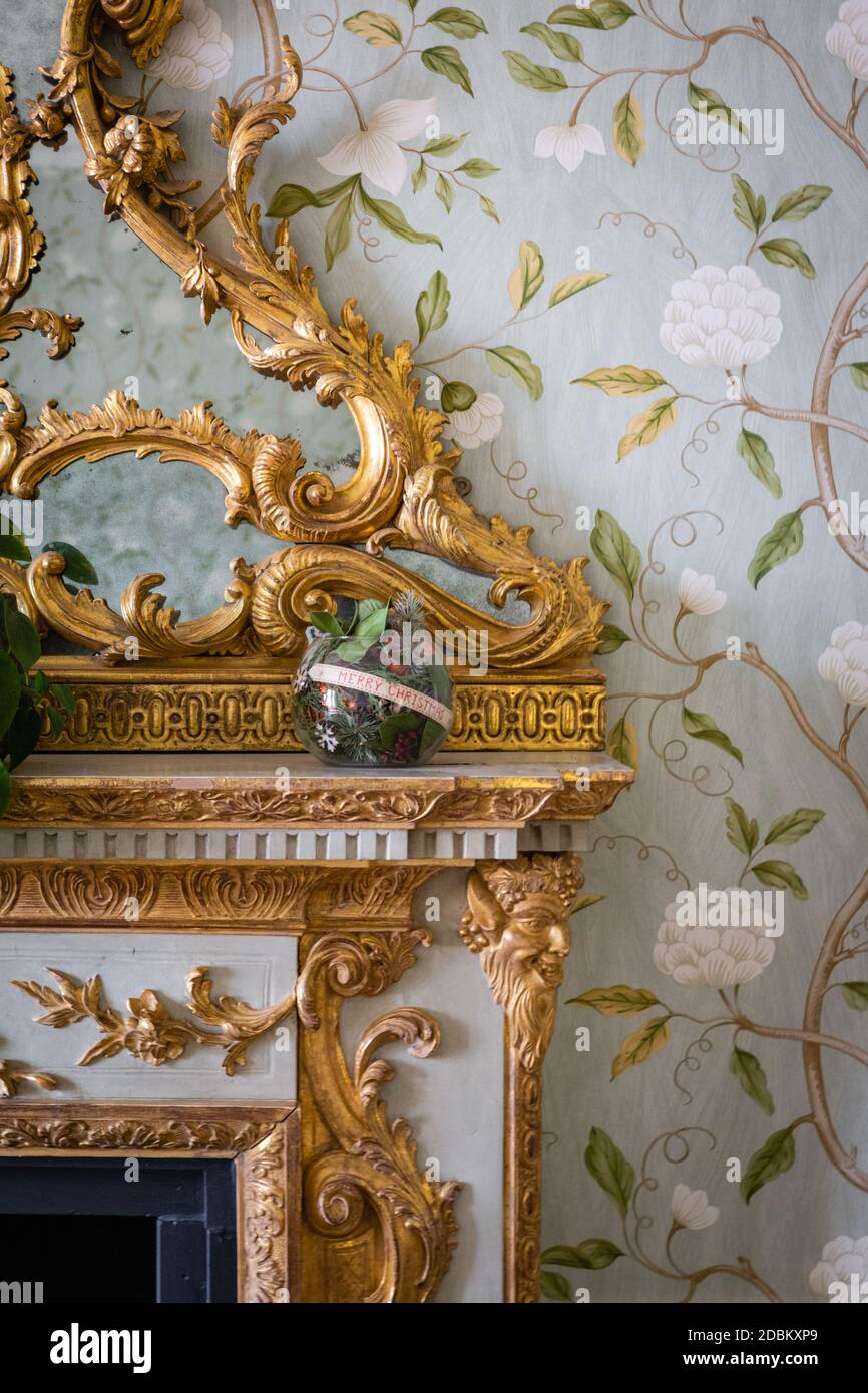 Une boule de Noël se trouve sur le manteau d'un feu orné de détails dorés complexes et d'un papier peint à fleurs. Banque D'Images
