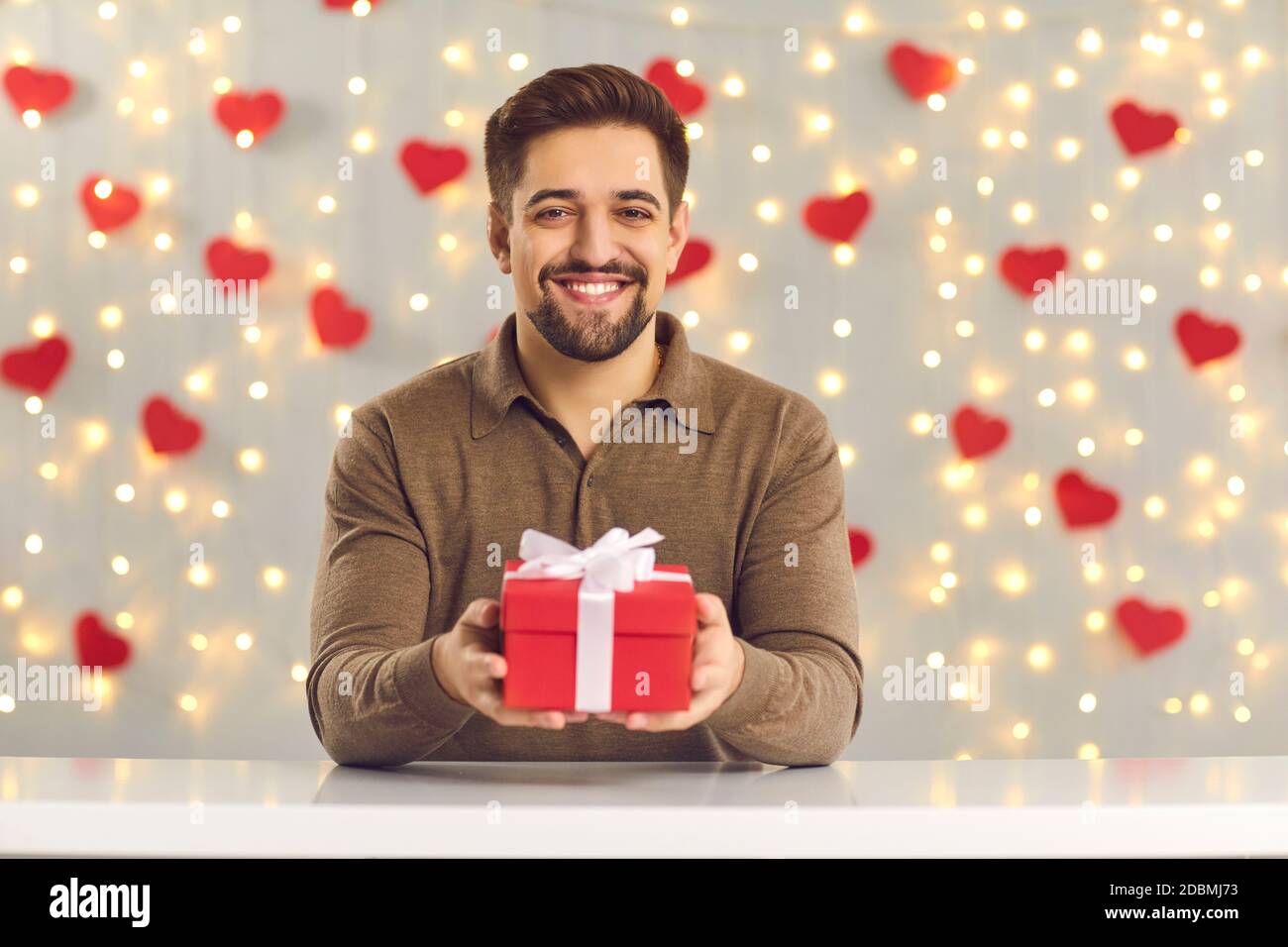 Jeune homme souriant tenant Saint Valentin présent assis à table contre décoration murale coeurs et lumières Banque D'Images