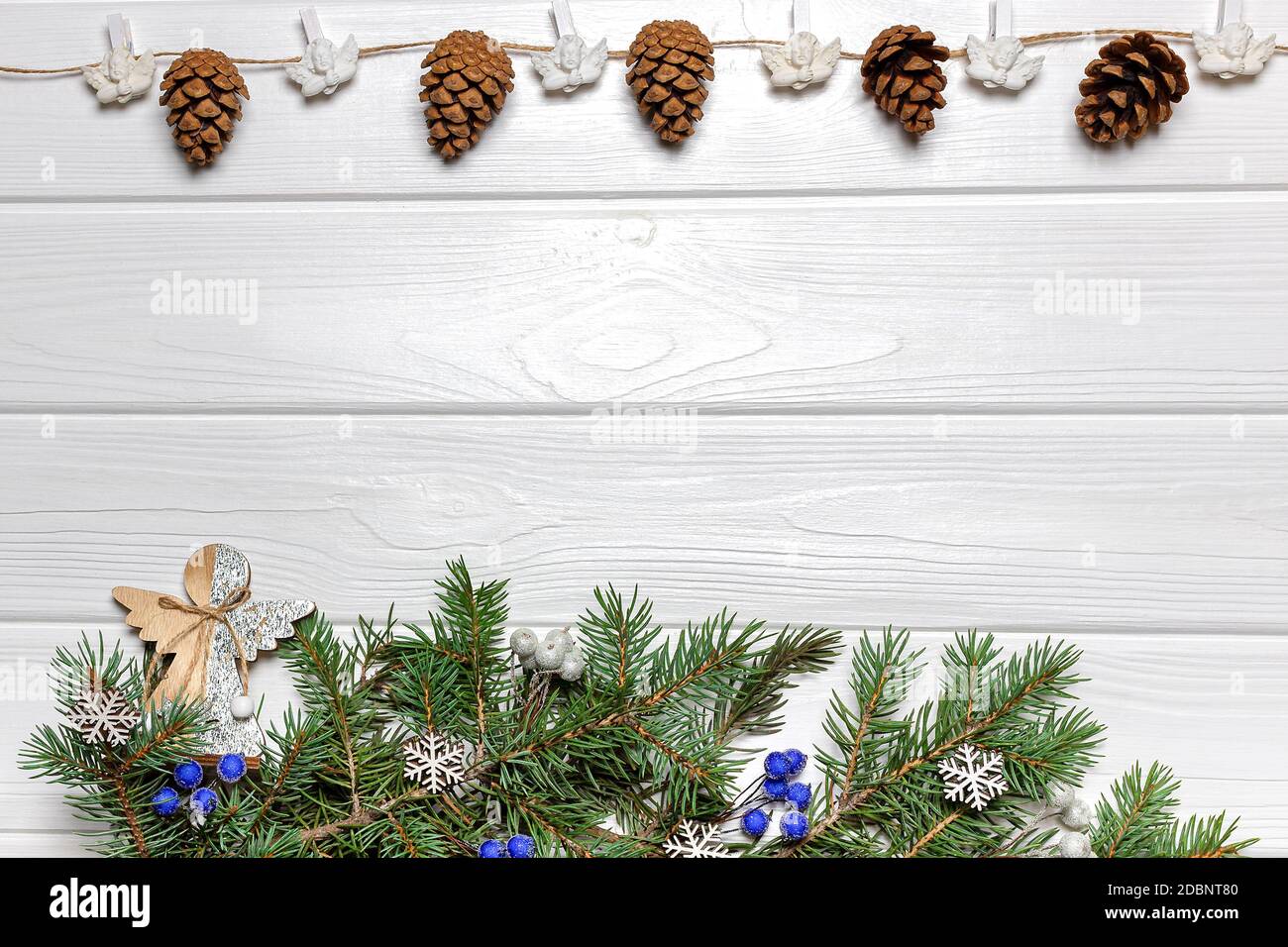 Guirlande avec des cônes et des anges, branches de pin avec ornements en bois et baies bleues sur fond de bois blanc. Modèle de carte de nouvelle année, espace de copie Banque D'Images