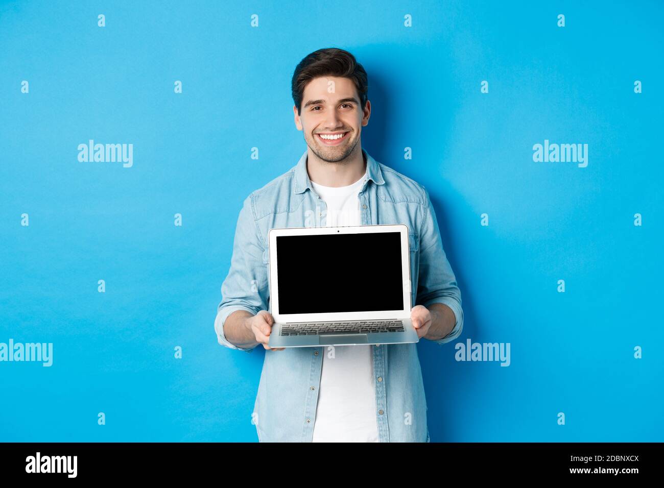Beau jeune homme présenter le produit sur l'écran d'ordinateur portable, montrant l'ordinateur et souriant, debout sur fond bleu Banque D'Images