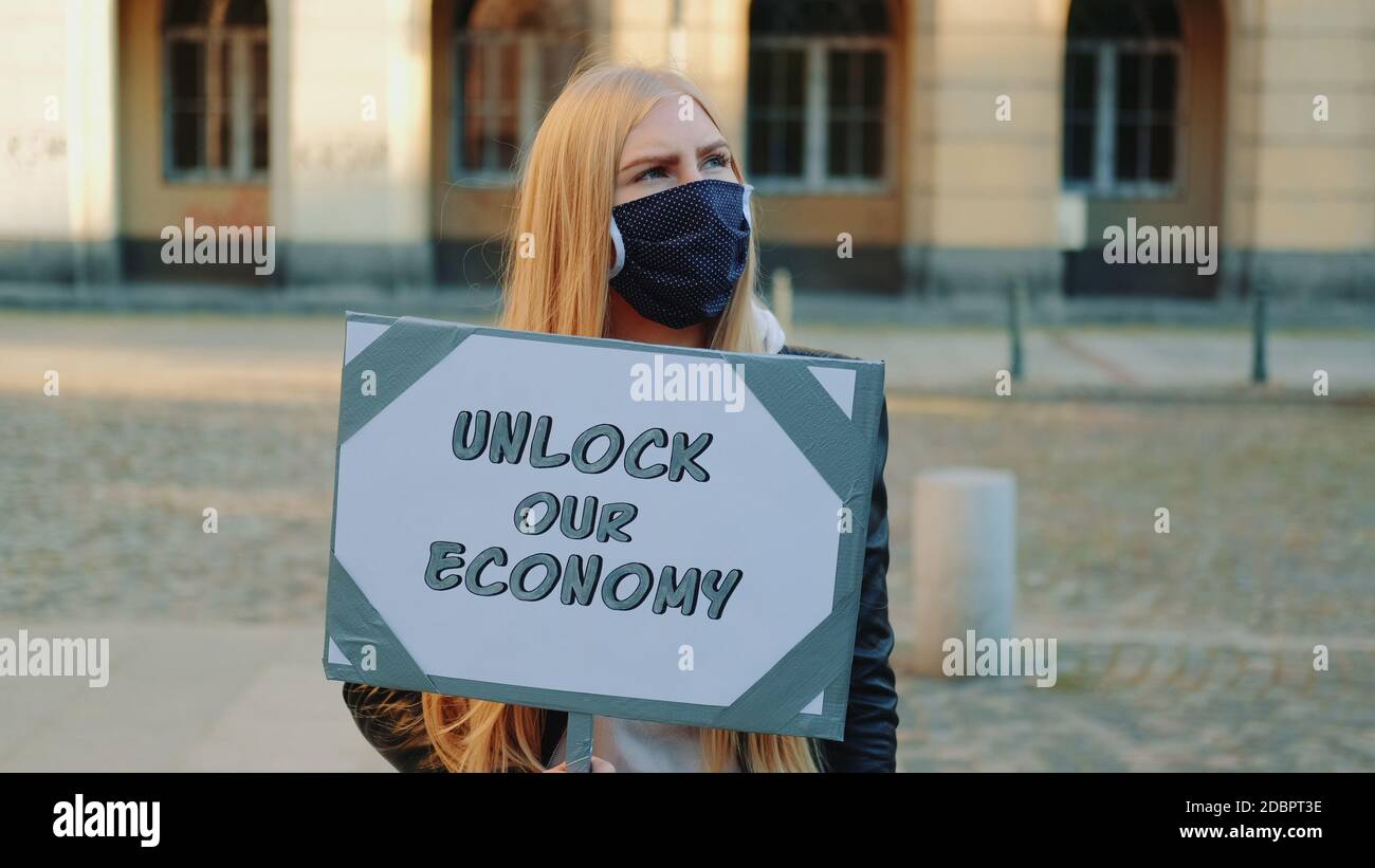 Jeune femme portant un masque médical appelant à déverrouiller l'économie en tenant le défroisseur. Promenade de protestation dans le centre-ville. Banque D'Images