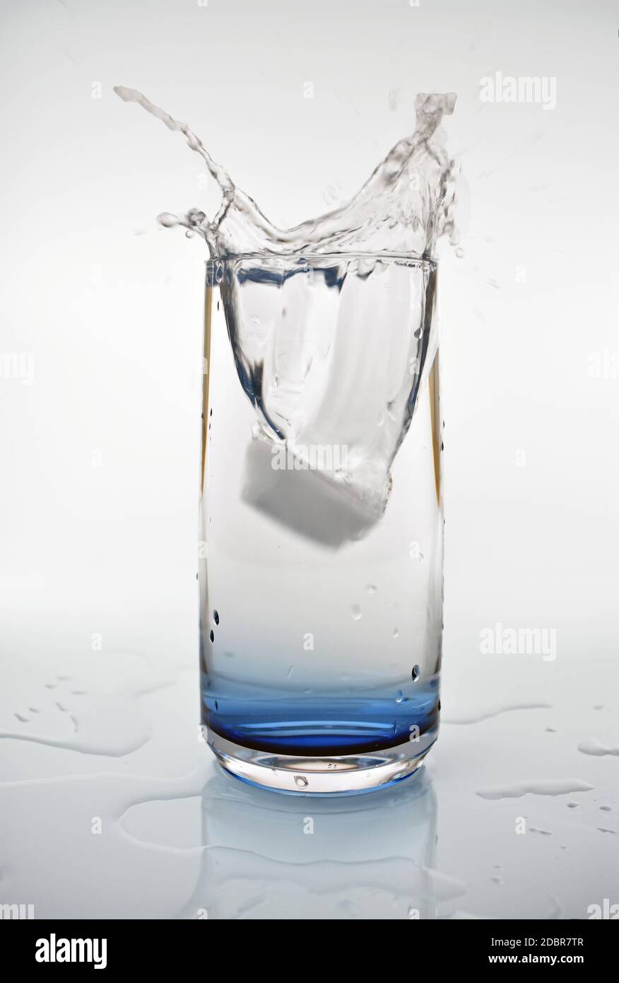 Un cube de glace tombant dans un verre d'eau créant une éclaboussure. Le verre teinté bleu se reflète dans les éclaboussures d'eau sous la boisson. Banque D'Images