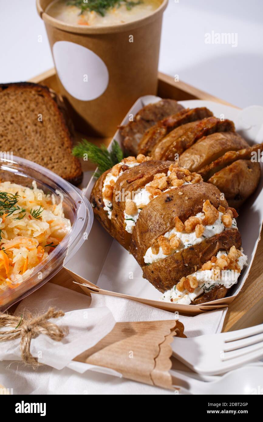 Vaisselle jetable, soupe dans une tasse de papier, pain, salade et pommes de terre cuites au four avec fromage cottage et noix sur un plateau en bois sur fond blanc. Blure Banque D'Images