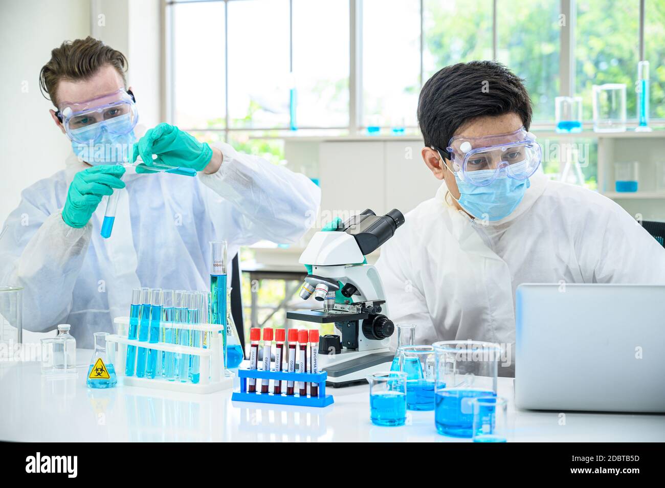 Couple de scientifiques mâles portant une combinaison de protection travaillant avec le microscope et Ordinateur portable et de nombreux équipements de laboratoire pour la recherche du vaccin contre le coronavirus à Banque D'Images