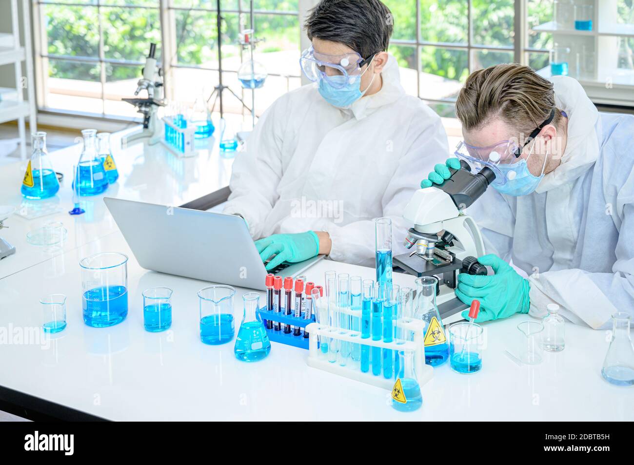 Couple de scientifiques mâles portant une combinaison de protection travaillant avec le microscope et Ordinateur portable et de nombreux équipements de laboratoire pour la recherche du vaccin contre le coronavirus à Banque D'Images