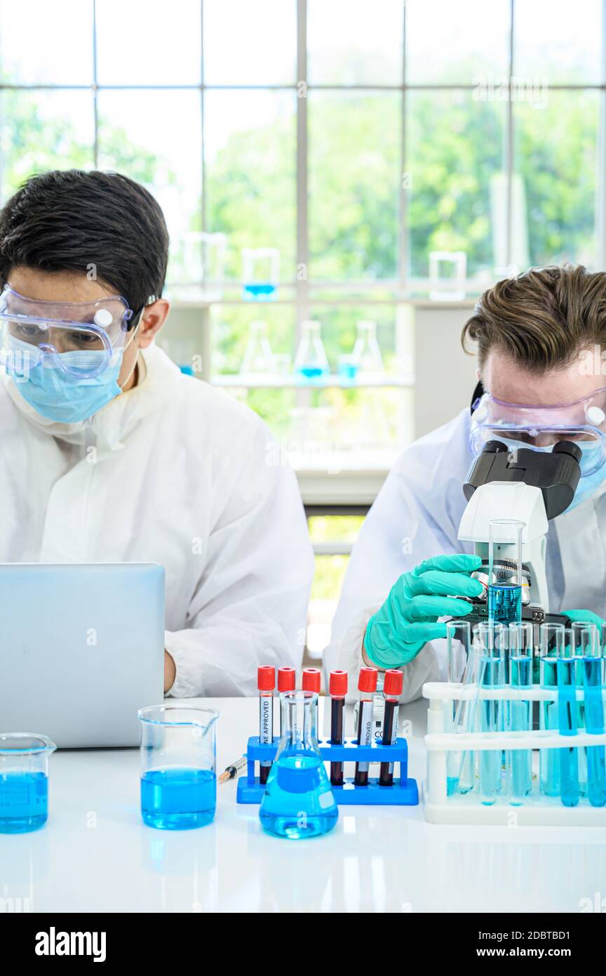 Couple de scientifiques mâles portant une combinaison de protection travaillant avec le microscope et Ordinateur portable et de nombreux équipements de laboratoire pour la recherche du vaccin contre le coronavirus à Banque D'Images