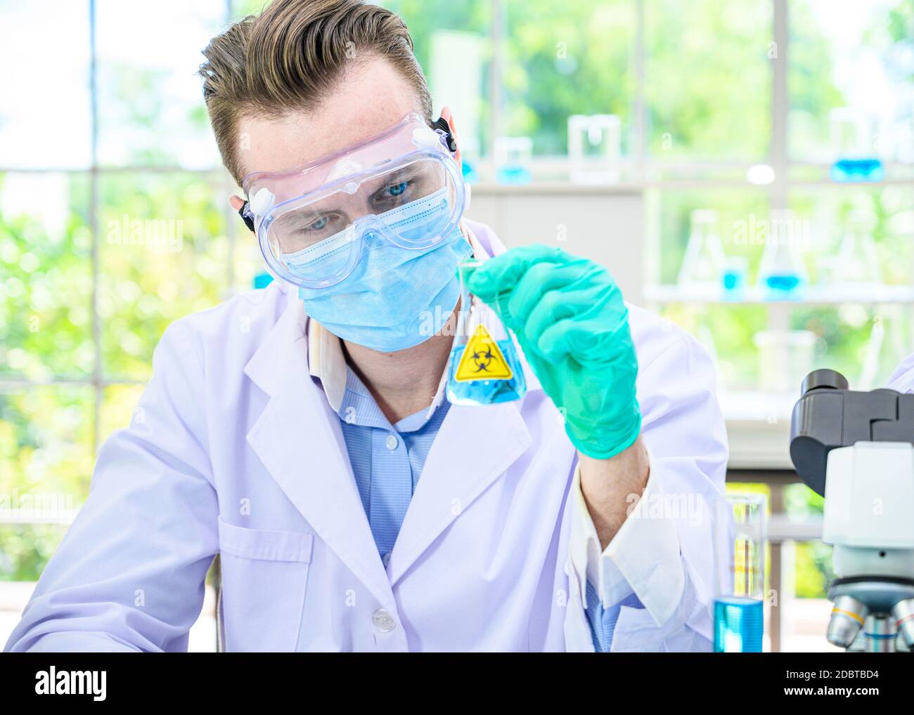 Un scientifique mâle porte une combinaison de protection pour le maintien d'un bécher ou d'un tube avec prélever du liquide à l'intérieur et travailler avec de nombreux équipements de laboratoire pour recherche en laboratoire Banque D'Images