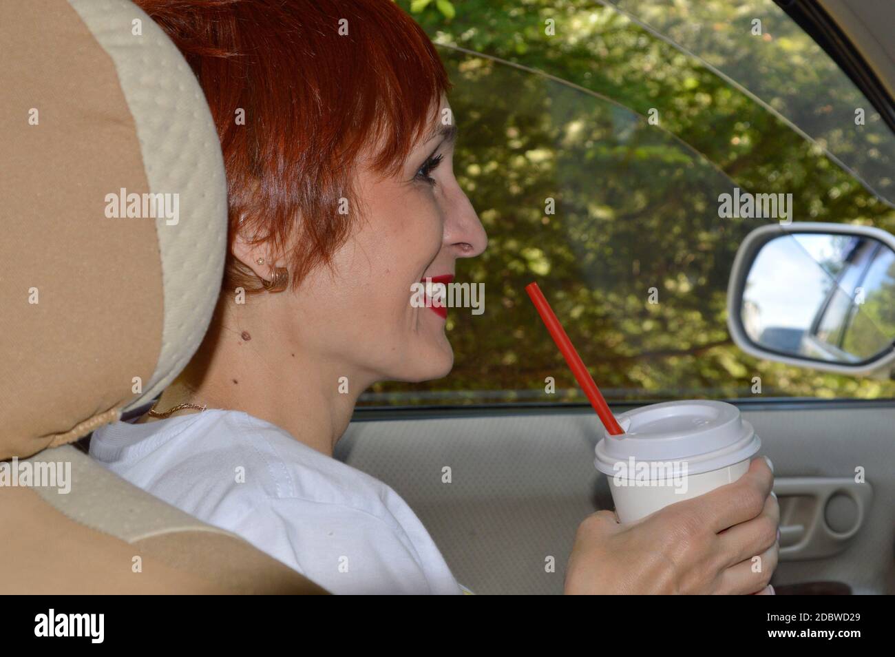 Jeune femme gaie passager avec cheveux rouges assis dans la voiture, tenant une tasse de plastique de café et souriant avec les dents. Aventures d'été Banque D'Images