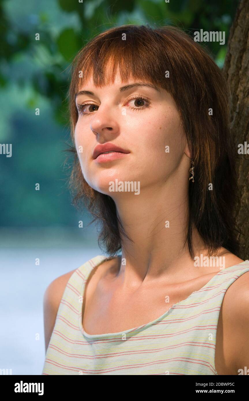 Portrait latéral de la tête et de l'épaule d'une jeune femme brunette se penchant contre un arbre, regardant sérieusement la caméra. Banque D'Images