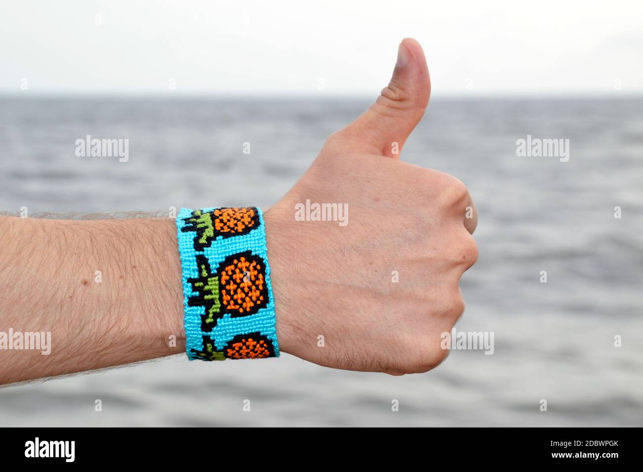 Bracelet d'amitié tissé bleu avec motif ananas sur les mâles main contre fond de mer. Signal du pouce Banque D'Images