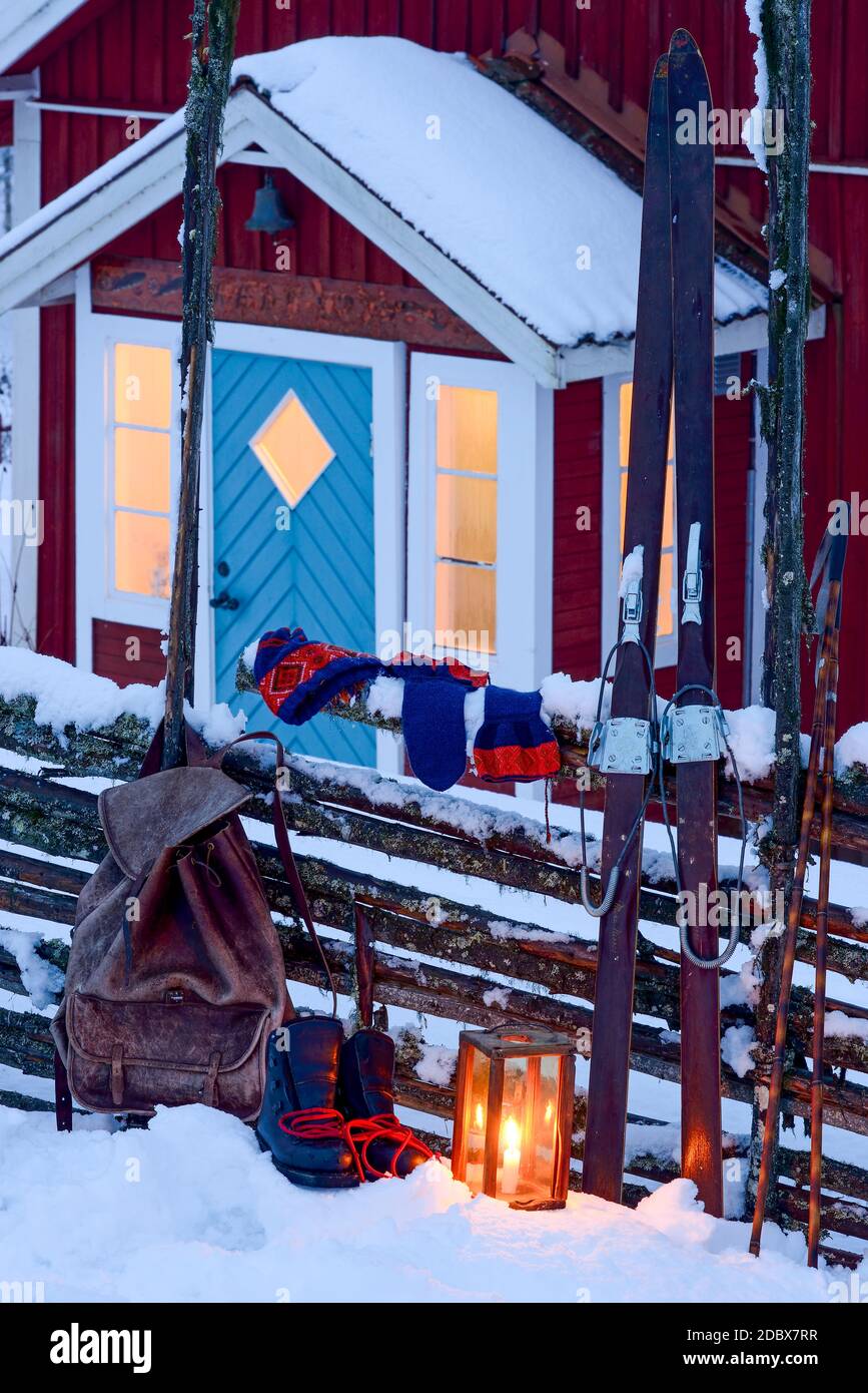 Une lanterne, un sac à dos, une paire de bateaux, une mitaine et une paire de cieux penchée sur une clôture devant un porche à une maison rouge suédoise traditionnelle en hiver Banque D'Images