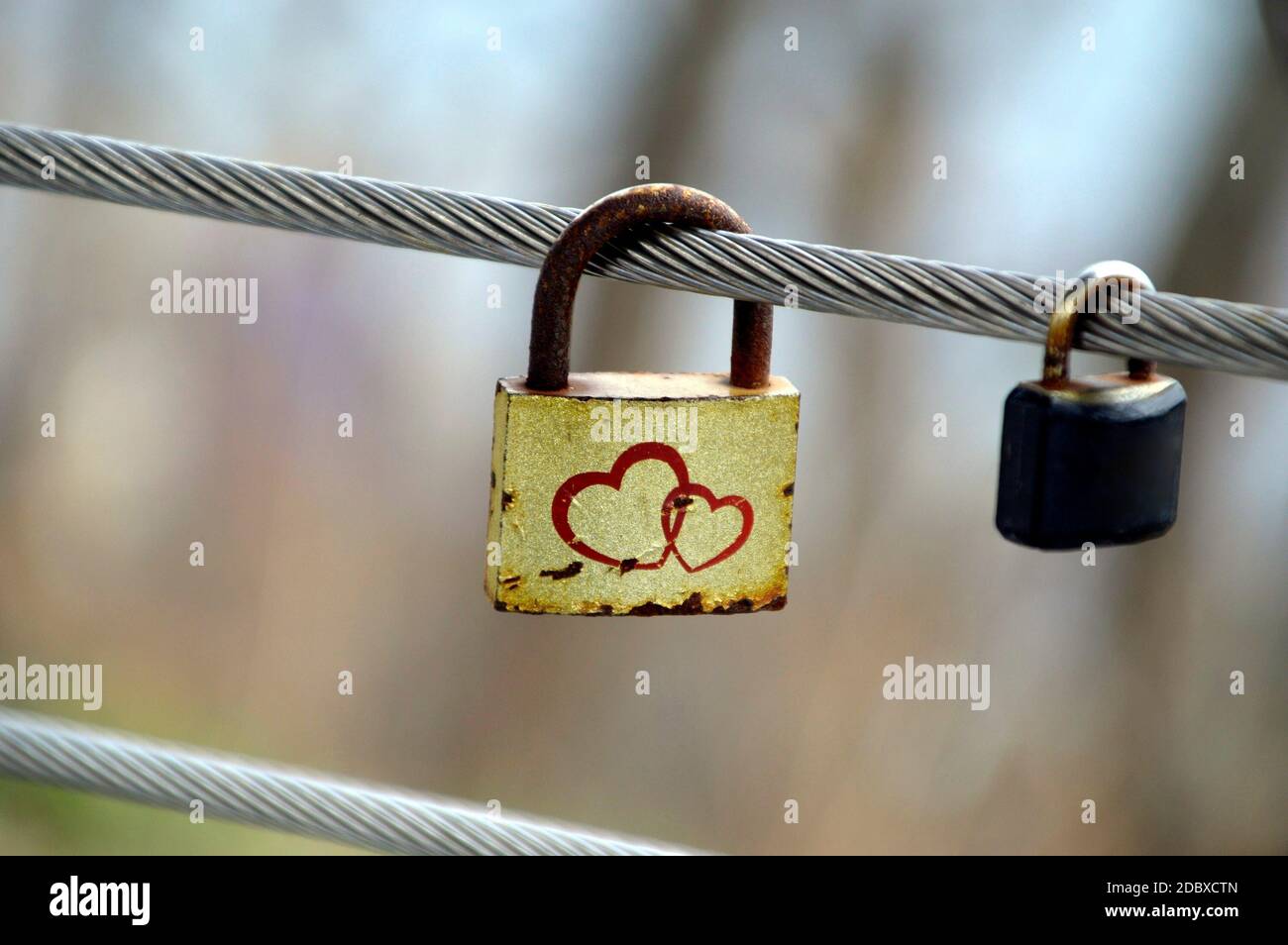 Vieux cadenas jaune rouillé avec deux coeurs accrochés sur un clôture en métal à côté d'un petit cadenas habituel à l'extérieur Banque D'Images