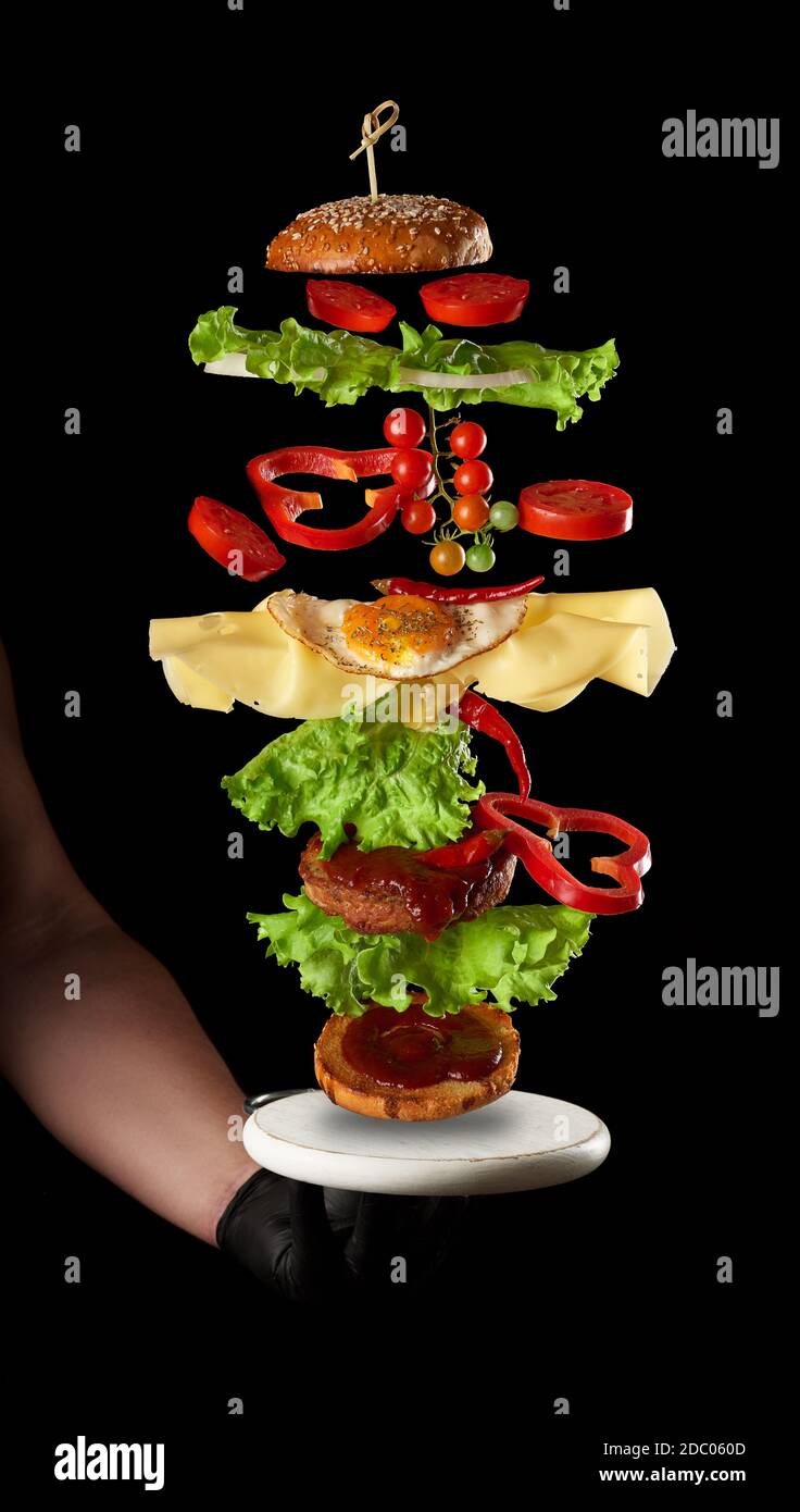 ingrédients de cheeseburger volant : côtelette de viande, pain de sésame, fromage, œuf frit, tomate et laitue verte sur fond noir, main d'un homme dans un g noir Banque D'Images