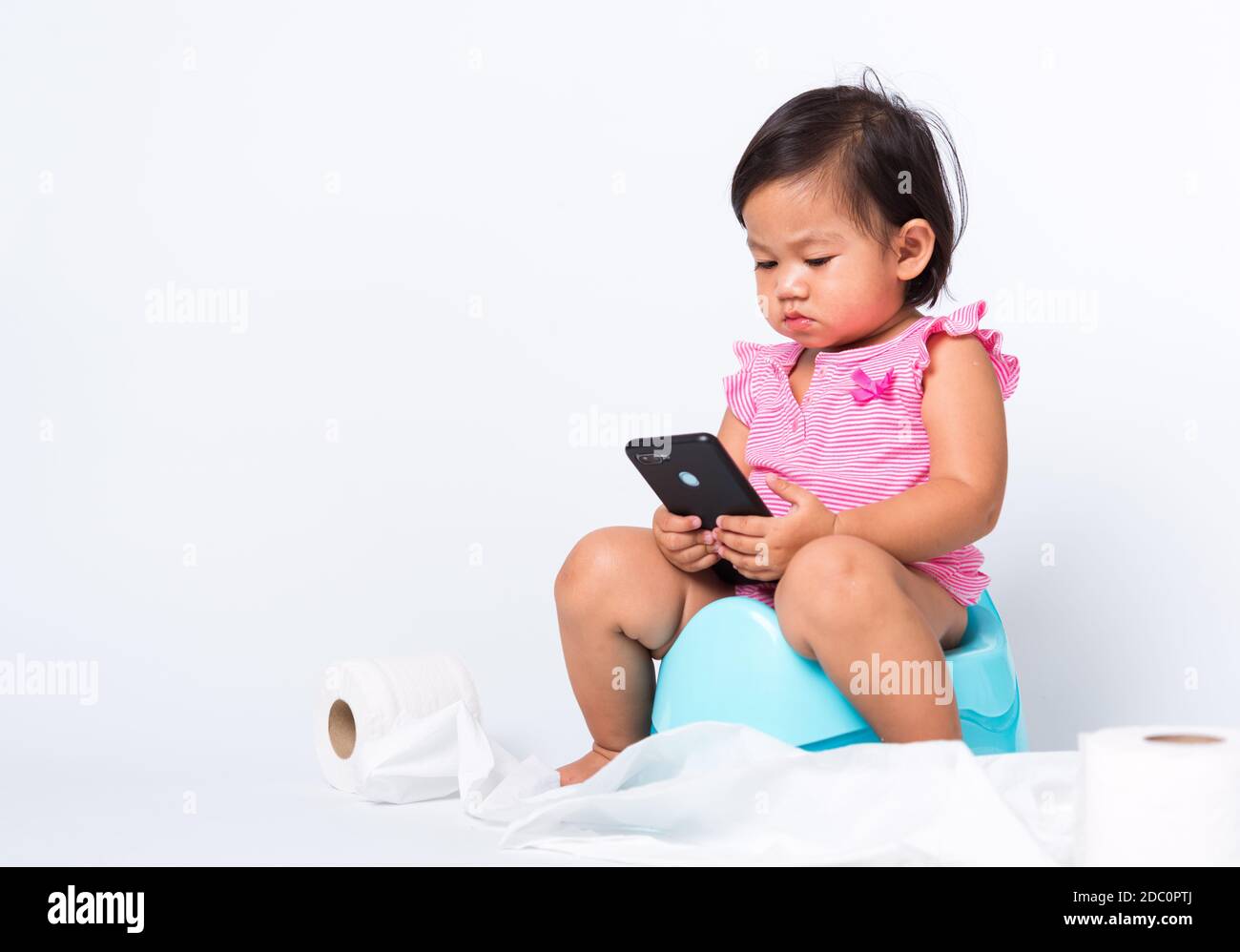 Asiatique petite mignonne bébé fille éducation entraînement à s'asseoir sur pot bleu de chambre ou pot et jouer smartphone mobile avec des rouleaux de papier de toilette, goujon Banque D'Images