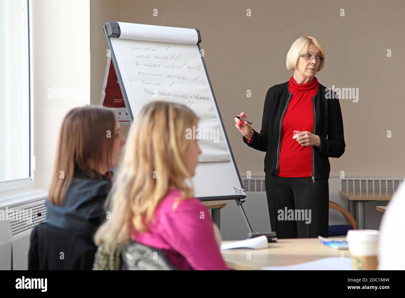 Une femme d'âge moyen mène une discussion de gestion - écrivant sur un tableau de conférence. Deux jeunes femmes s'assoient à un bureau au premier plan. Banque D'Images