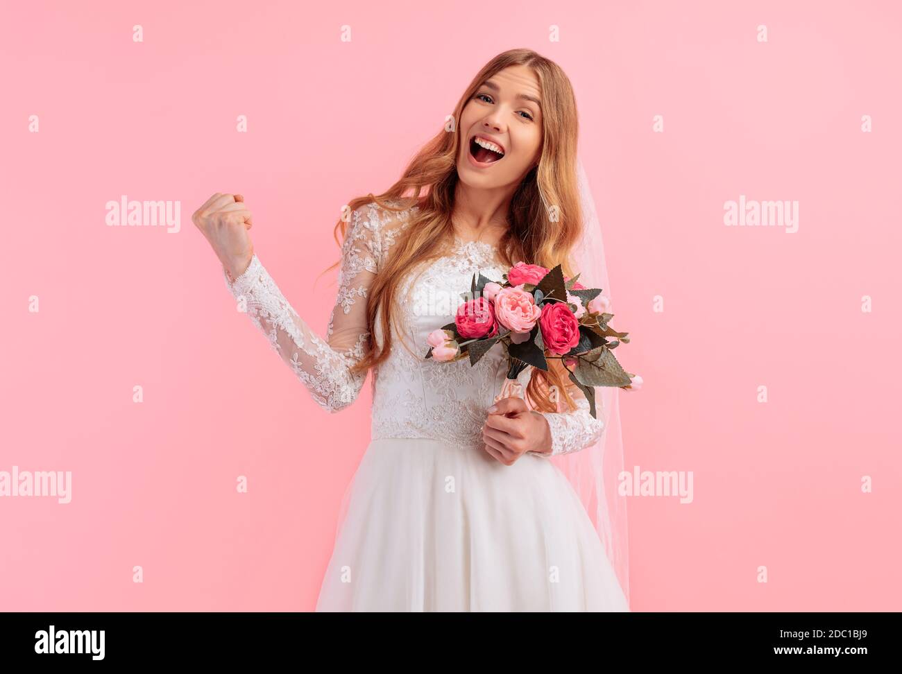 Une mariée heureuse et excitée avec un bouquet de fleurs dans une robe de mariage célèbre la victoire sur un fond rose. Mariage Banque D'Images