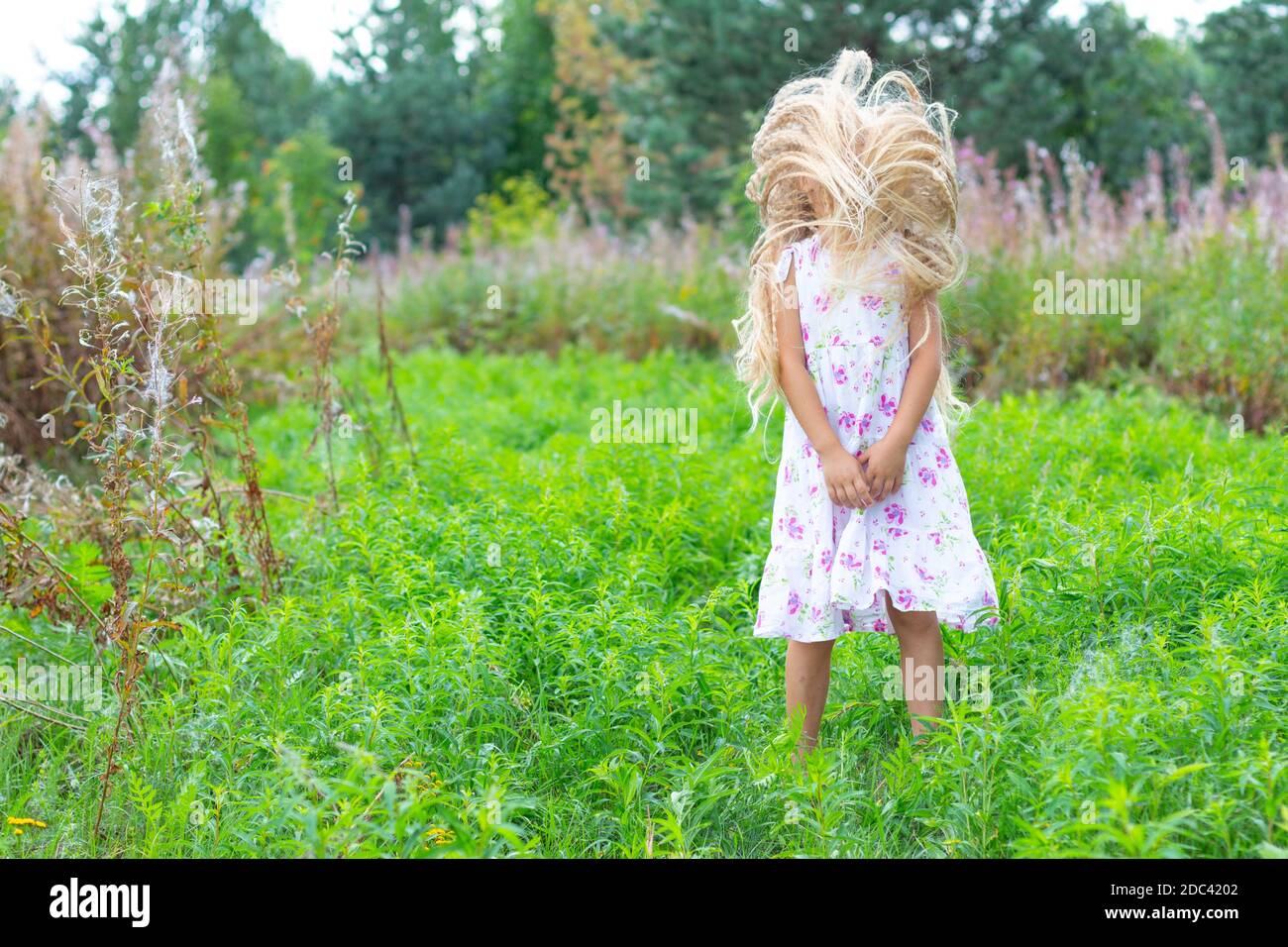 Une fille dans une sundress blanche, avec des cheveux blonds luxuriants, se tient dans un pré en fleurs, a secoué sa tête, aucun visage n'est visible, ses mains sont pliées. Banque D'Images