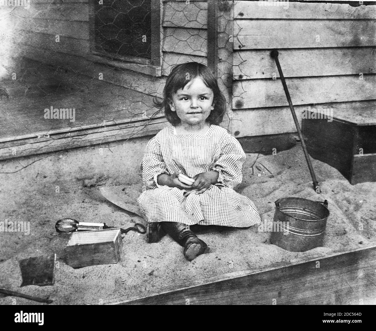 Voici une petite fille heureuse assise dans son bac à sable, vers 1910. Cette boîte privée est séparée du porche avant de sa maison par du fil de poulet, qui est cloué à la parement en bois de la maison. Sur la voie d'évitement, notez les taches de rouille où les clous sont coulés près du coin du bâtiment. La fille a le tire-paille de sa mère, un seau, et d'autres articles qu'un enfant pourrait trouver utiles dans un bac à sable. Pour voir mes enfants-liés des images vintage, recherche: Prestor vintage Kids Banque D'Images
