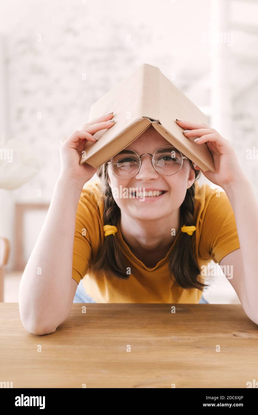 Jeune belle femme étudiante en t-shirt orange et lunettes lit le livre assis à la table. Préparation aux examens, étude à domicile sur l'auto-isolement Banque D'Images