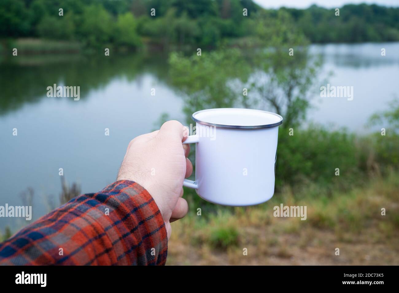 un homme avec une chemise de bûcherons tenant une tasse en émail devant un lac tout en randonnée et en buvant du café ou du thé frais Banque D'Images