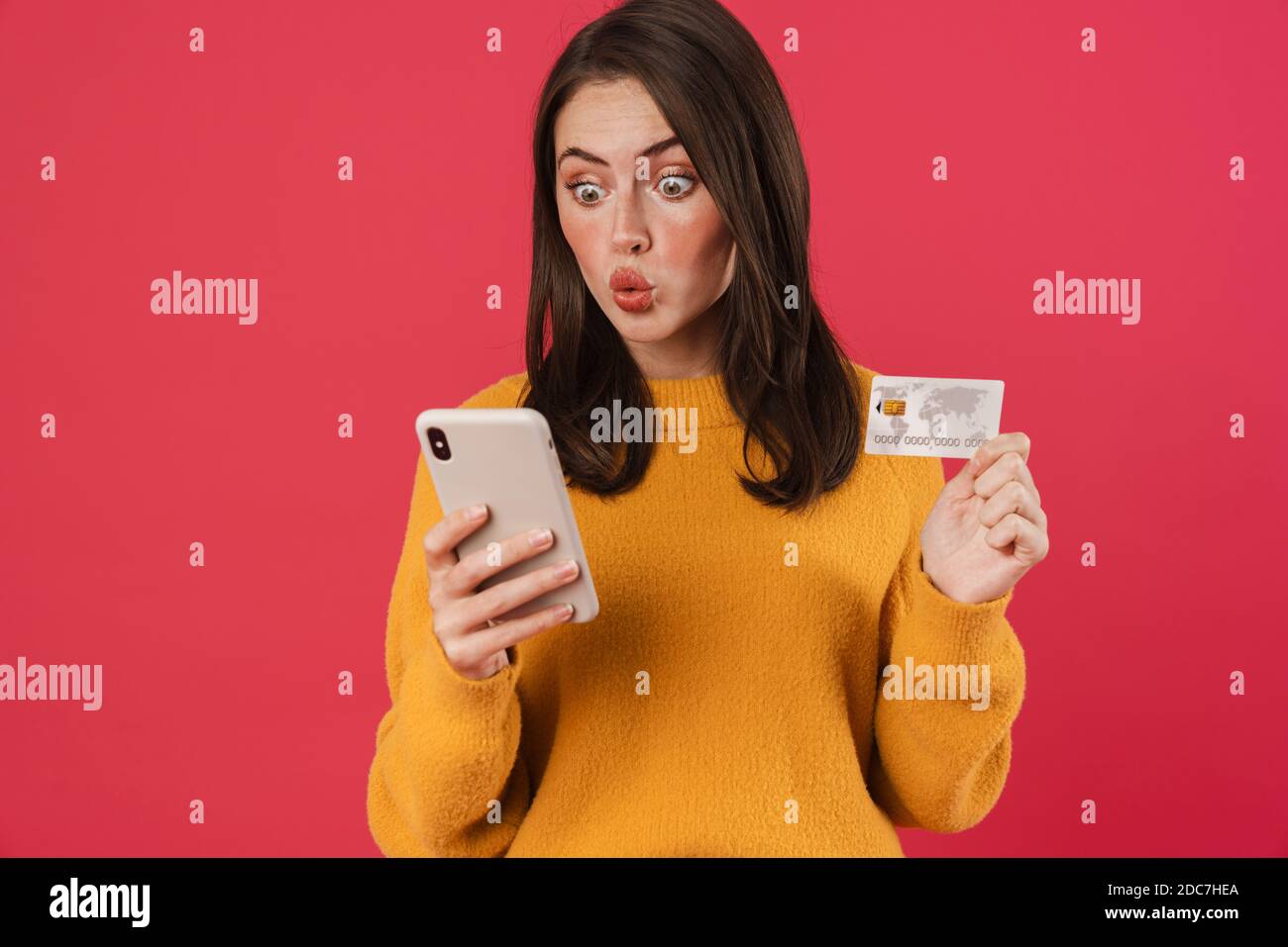 Image d'une jeune fille caucasienne choquée posant avec un téléphone portable et un crédit carte isolée sur fond rose Banque D'Images
