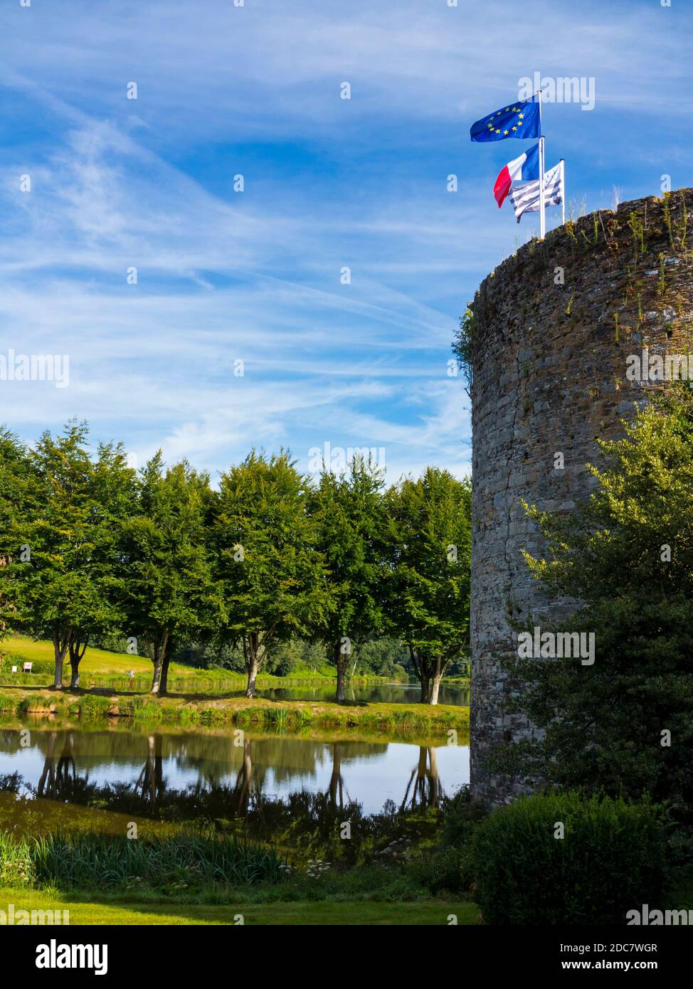 Tour en pierre et avenue d'arbres se reflétant dans l'eau au Haut-Corlay, dans l'ouest de la Bretagne, en France, avec des drapeaux français et de l'Union européenne volant sur un poteau. Banque D'Images