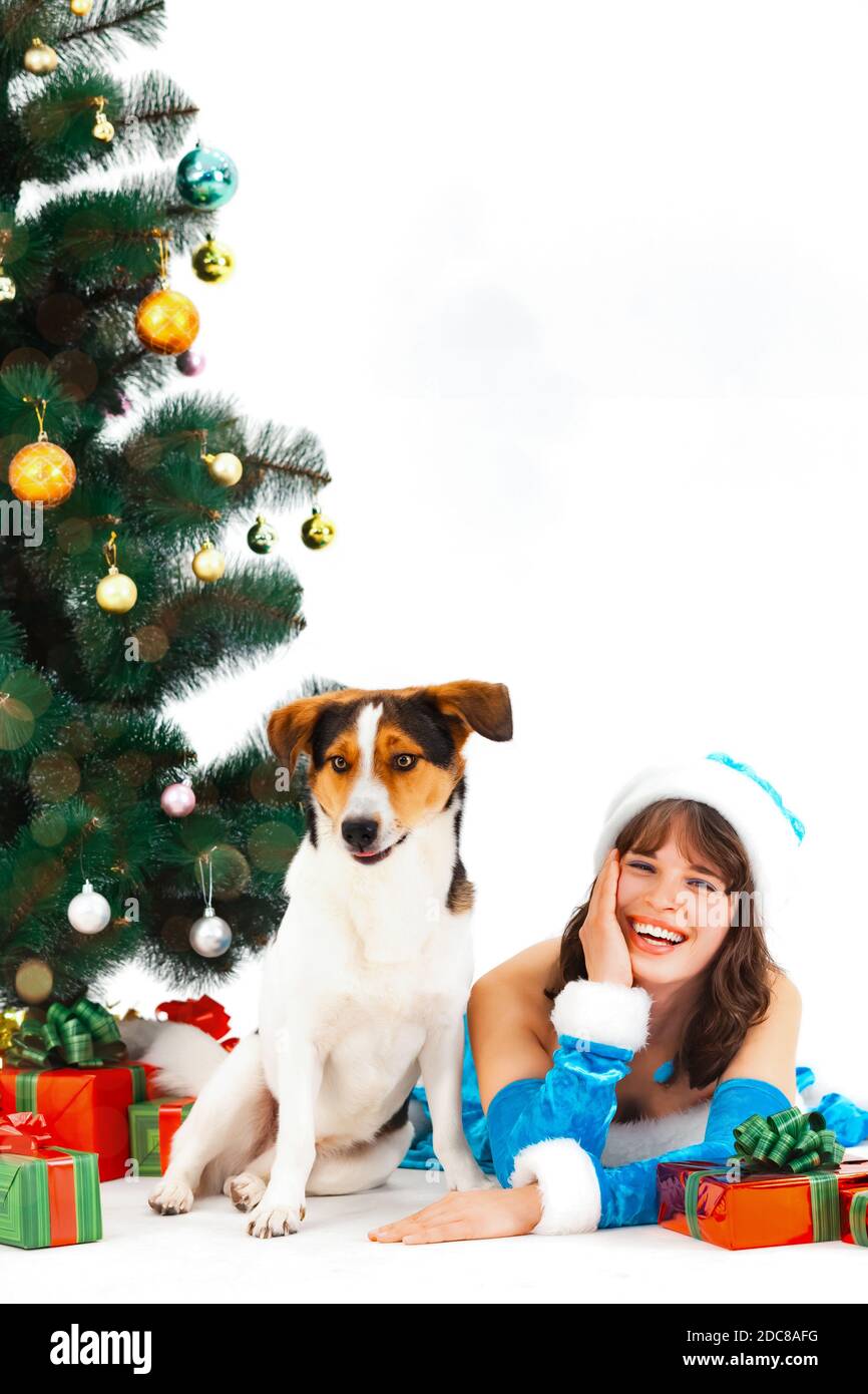 Une jeune femme en costume de père noël est en train de mentir et de rire avec un chien sur un fond blanc à côté d'un Arbre de Noël et cadeaux Banque D'Images