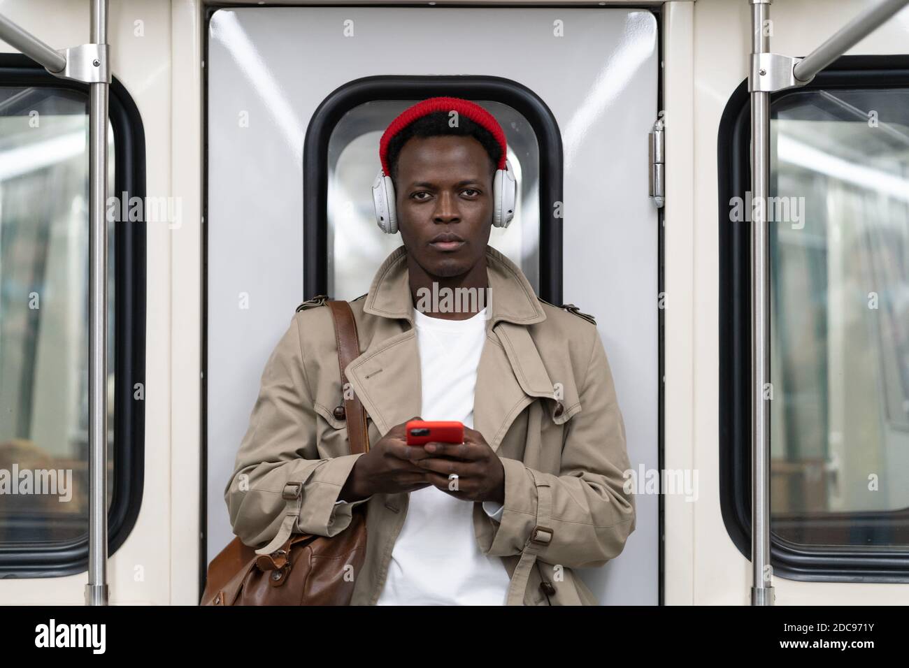 Homme millénaire afro-américain en chapeau rouge, stand de trench coat dans le métro, à l'aide d'un téléphone mobile, écoute de la musique avec des écouteurs sans fil dans le TR public Banque D'Images