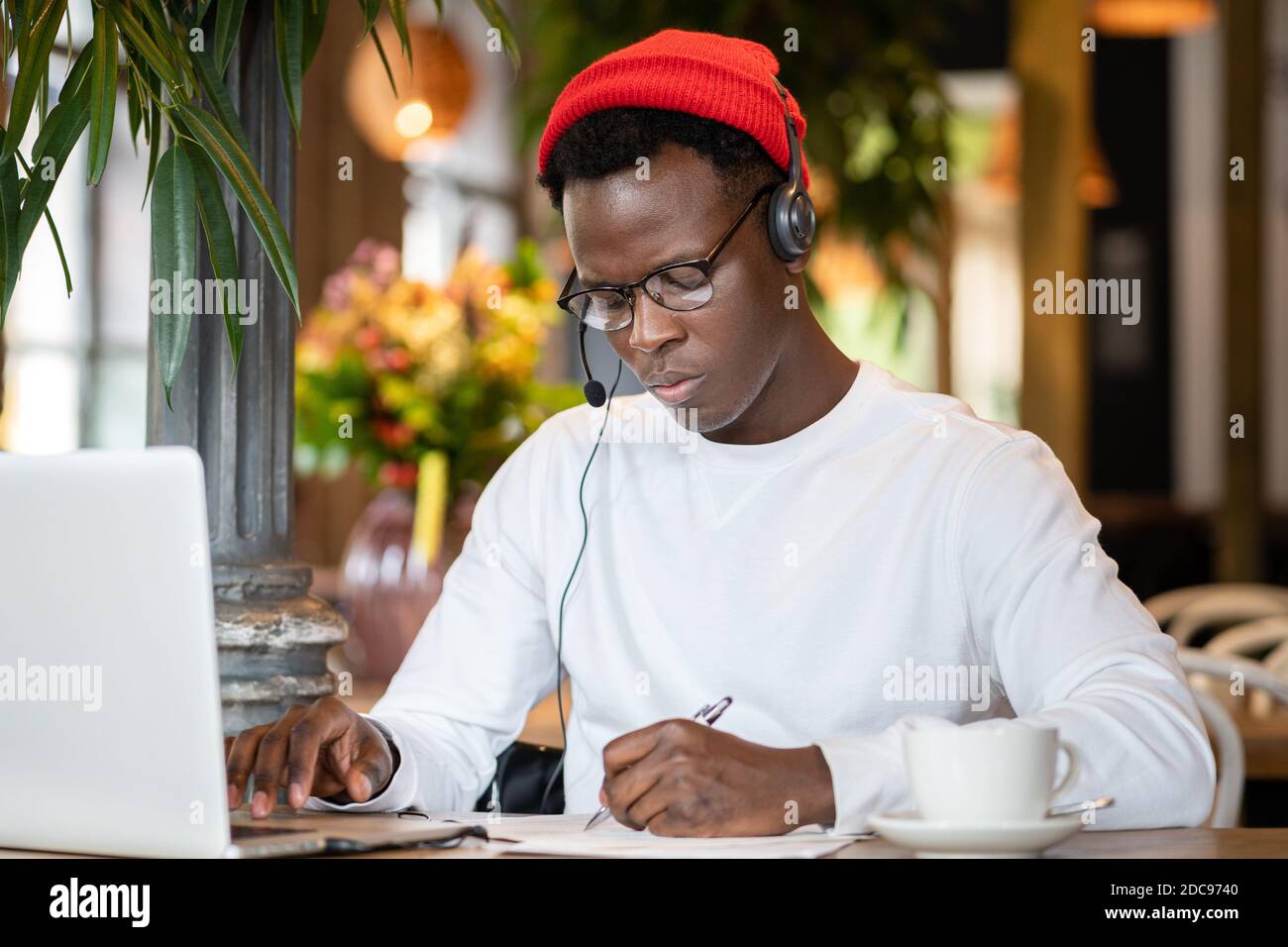 Homme noir millénaire concentré dans un casque porter un chapeau rouge, regarder un séminaire en ligne éducatif sur un ordinateur portable, travailler en ligne à distance dans un café, écouter une leçon audio Banque D'Images