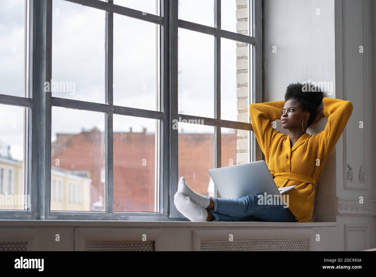 Femme millénaire afro-américaine décontractée avec une coiffure afro porter un gilet jaune, assis sur un rebord de fenêtre, se reposer, prendre une pause de travail sur un ordinateur portable, Banque D'Images