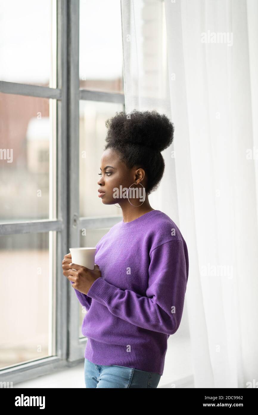Femme biraciale millénaire afro-américaine pensive avec bijoux tendance boucles d'oreilles porter un chandail violet pensant, regardant à travers la fenêtre, tenant le coup Banque D'Images