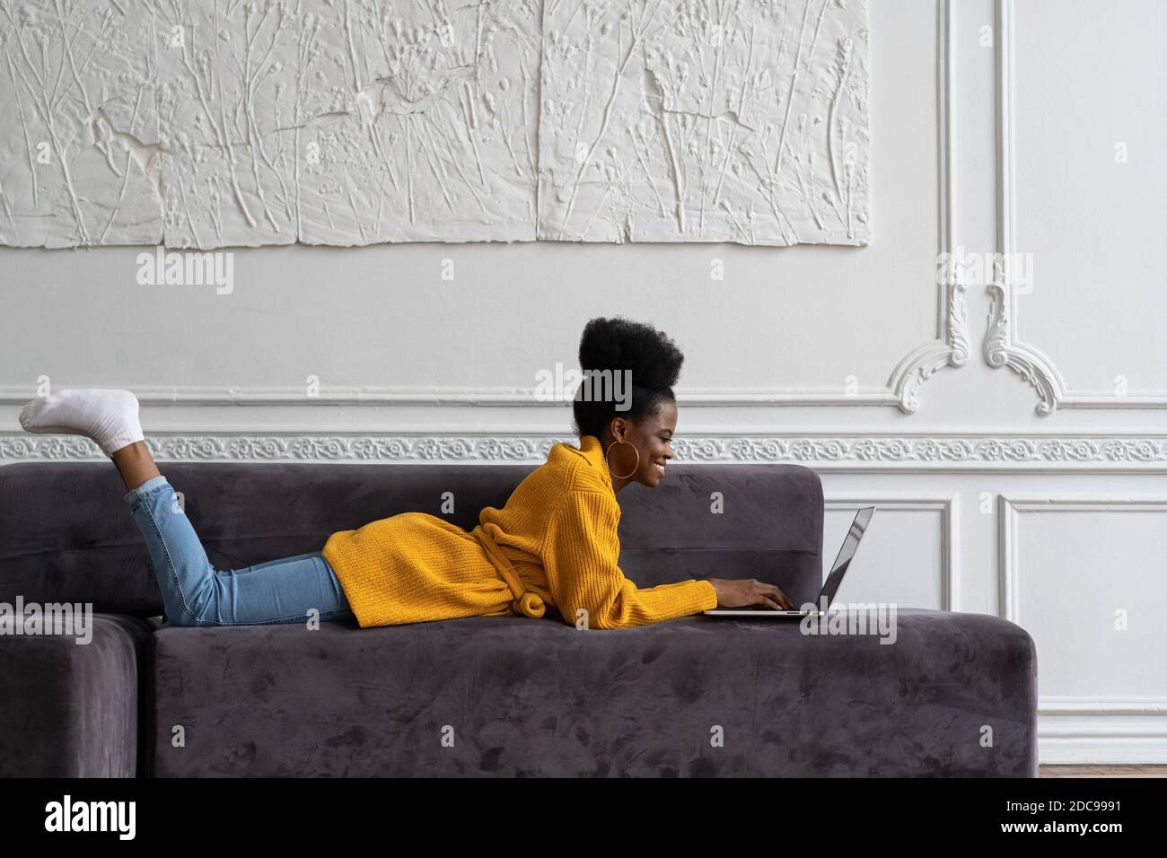 Souriante femme afro-américaine biraciale avec une coiffure afro-américaine dans un gilet jaune allongé sur un canapé, au repos, regardant la webcam de la caméra et parlant sur la vidéo cha Banque D'Images