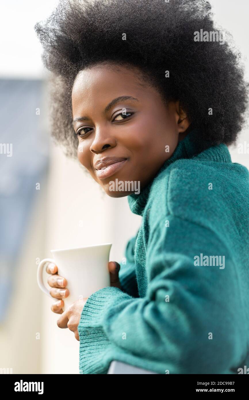 Adorable jeune femme biraciale afro-américaine appréciant une tasse de thé ou de café, porter un pull vert surdimensionné, regardant l'appareil photo, debout sur la ba Banque D'Images