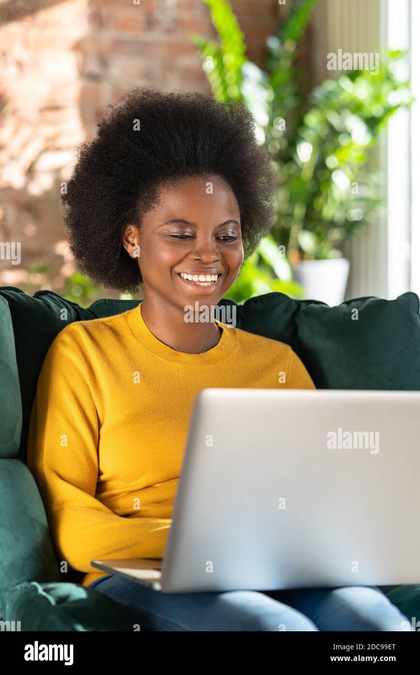 Jeune femme biracial afro-américaine souriante avec une coiffure afro-américaine en chandail jaune assis dans une chaise verte, travail à distance sur un ordinateur portable, chat et talkin Banque D'Images