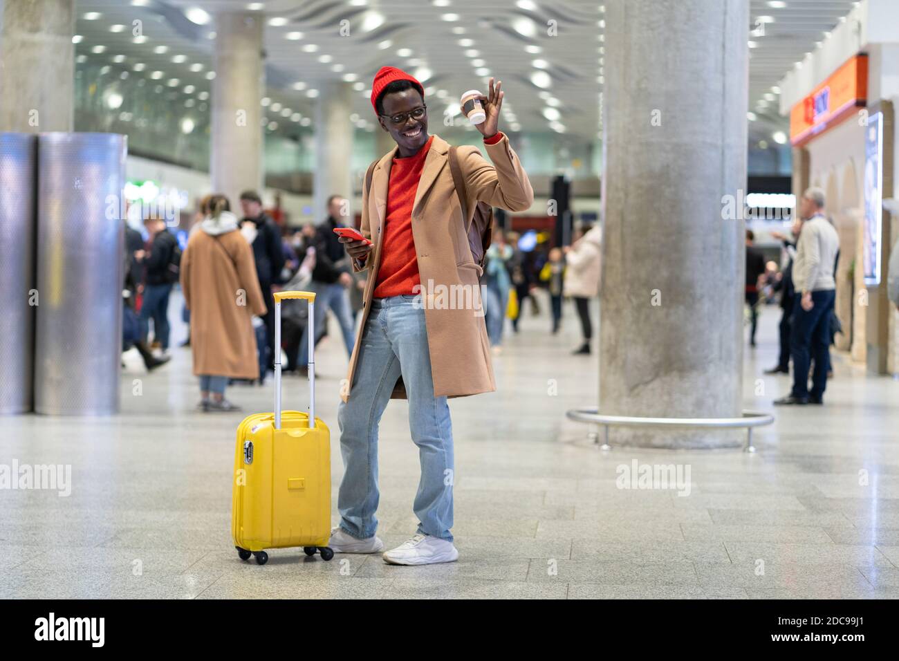 Voyageur afro-américain homme millénaire avec valise jaune dans le terminal de l'aéroport après l'arrivée, tenant le téléphone mobile et une tasse de papier de café, rai Banque D'Images