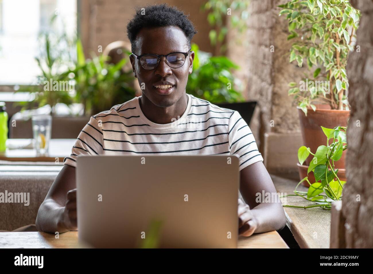 Jeune étudiant noir en t-shirt nu, en train de porter des lunettes, regarder un séminaire en ligne éducatif sur ordinateur portable, travailler en ligne à distance dans un café, étudier, préparer Banque D'Images