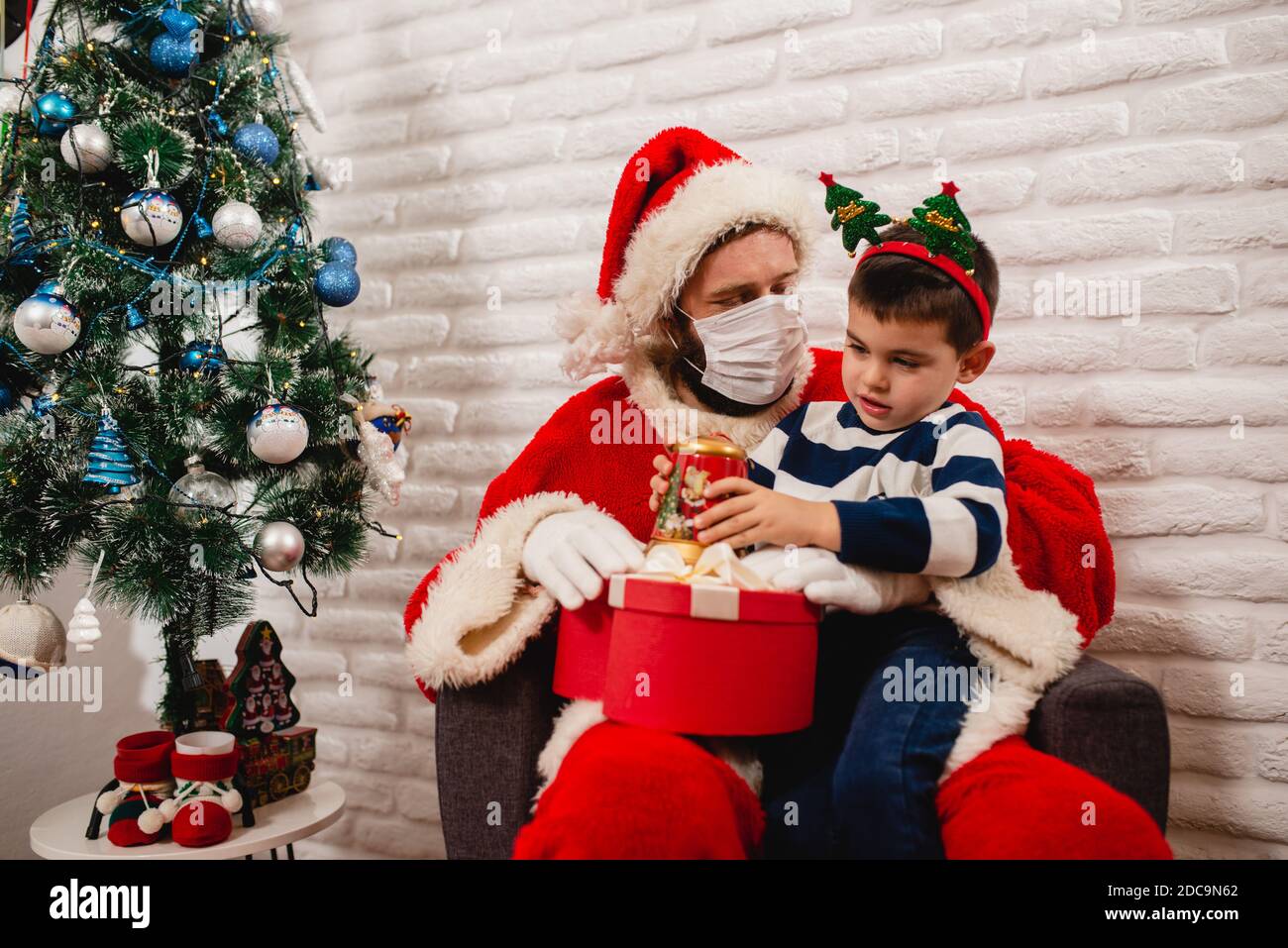 Un garçon de 5 ans excités avec une prop sur sa tête assis sur les genoux du Père Noël avec un masque facial et ouvrant un cadeau de Noël à côté de l'arbre de Noël. Hiver Banque D'Images